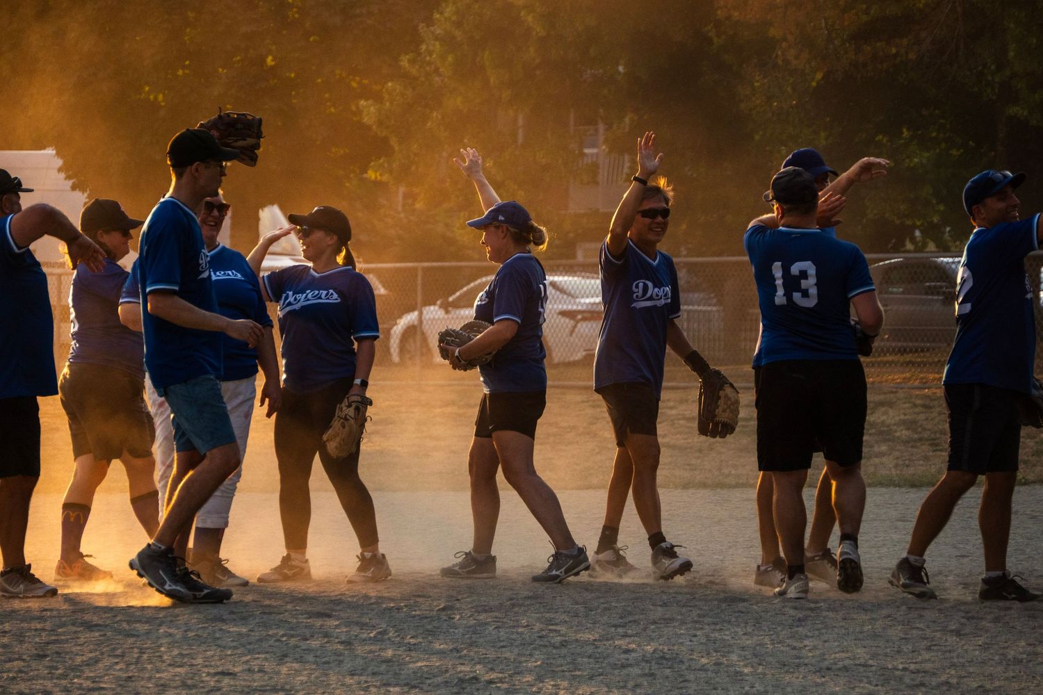 Softball teams exchange handshakes in dusty golden sunset light after a game.