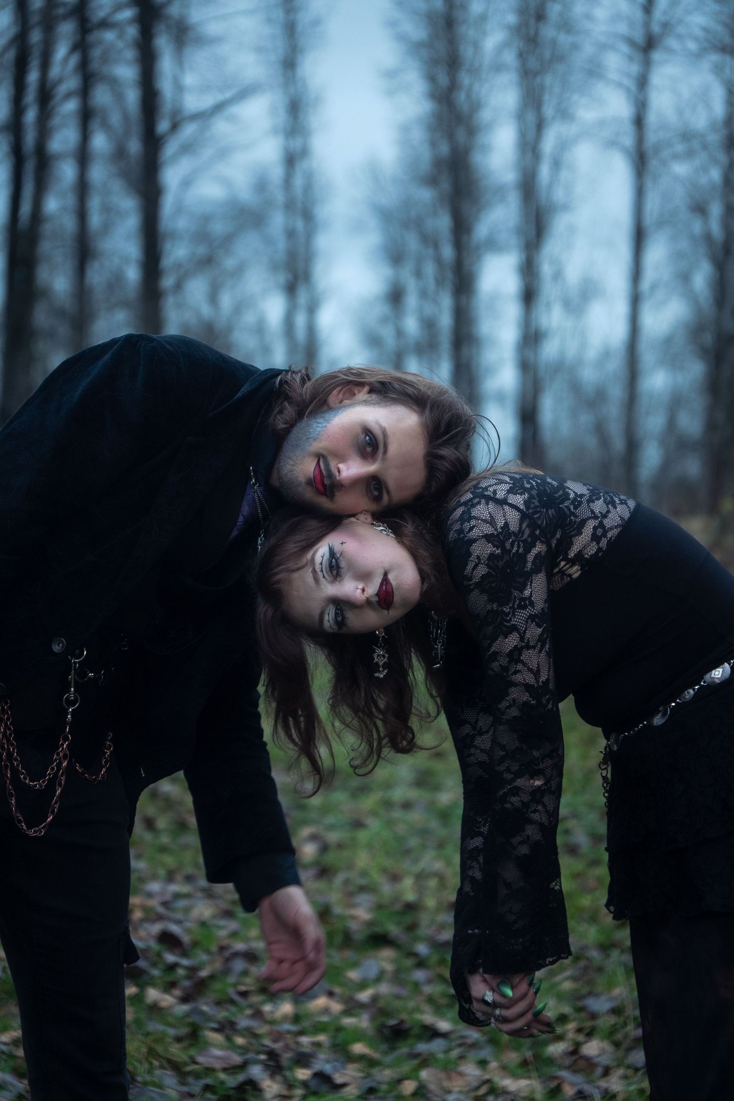 Two gothic individuals in black lean together cheek to cheek in a dark forest.