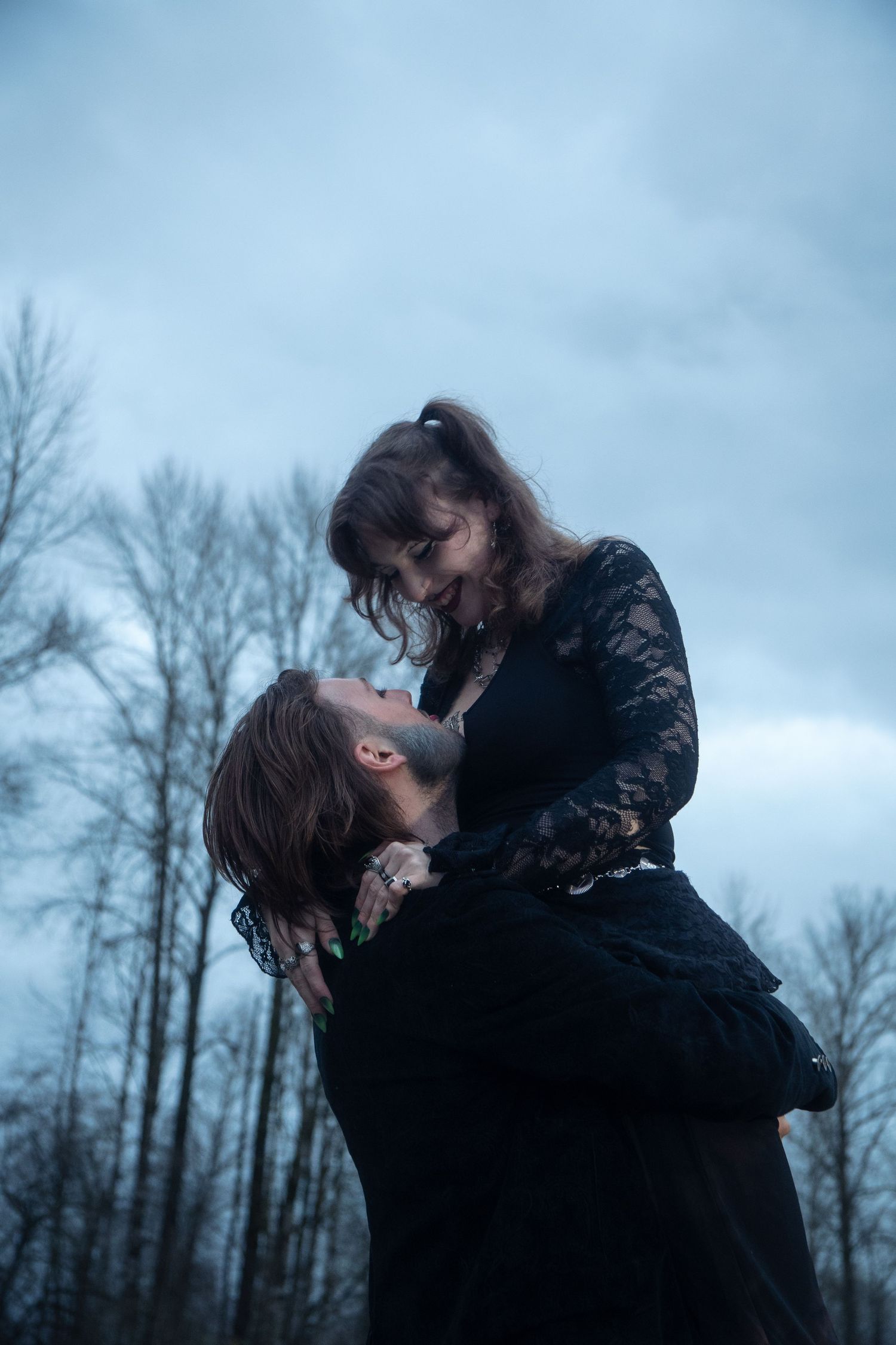 A man lifts a woman in a black lace dress as they share a romantic moment against a dramatic cloudy sky.