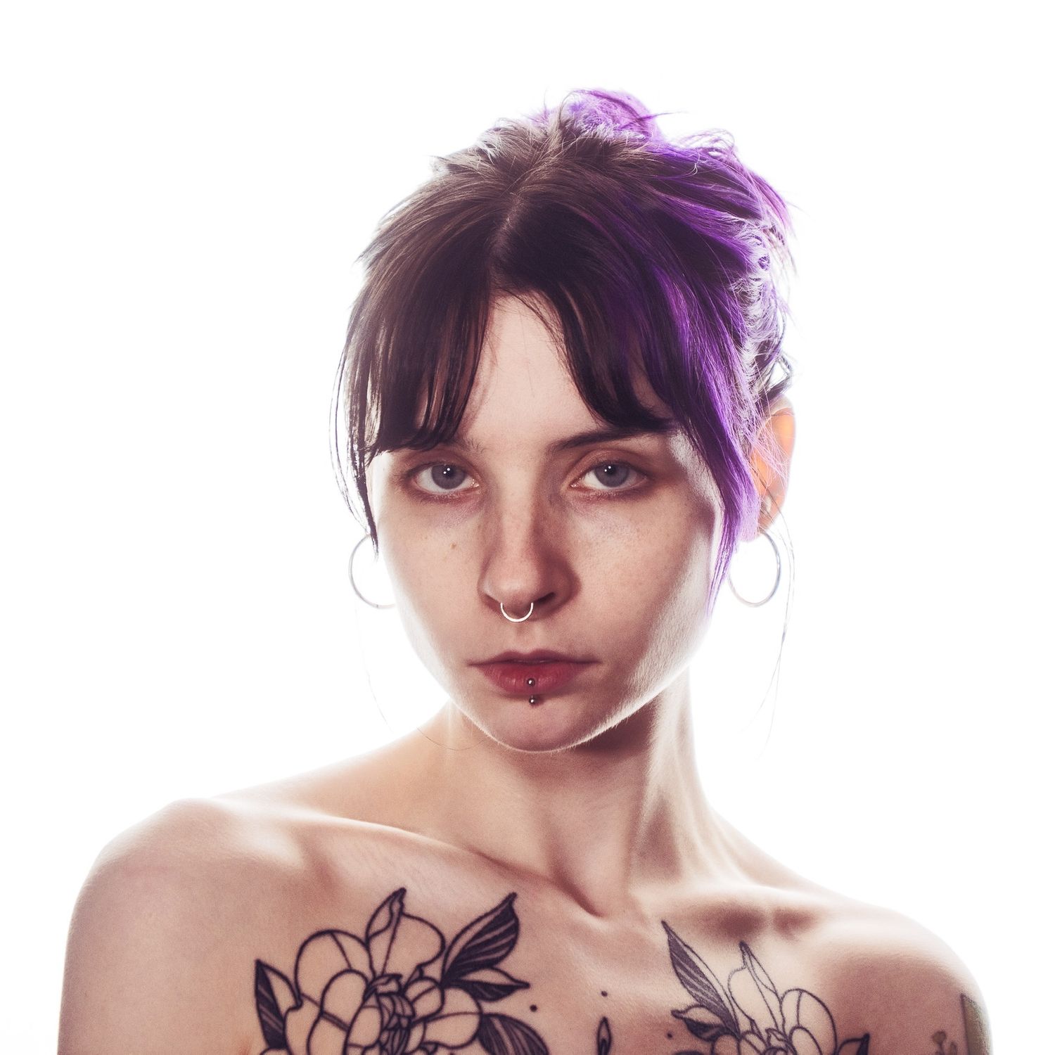 Woman with purple highlighted hair and floral chest tattoos wearing minimal clothing against white background.