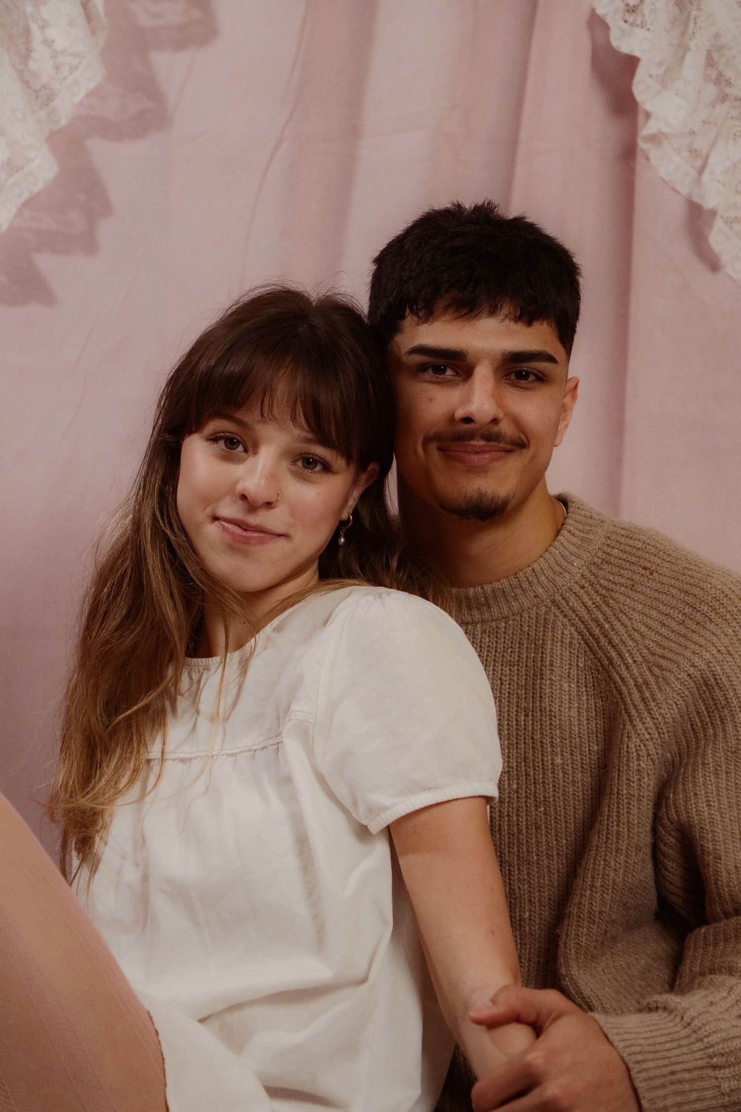 Young couple holding hands and smiling together against a soft pink lace curtain backdrop.