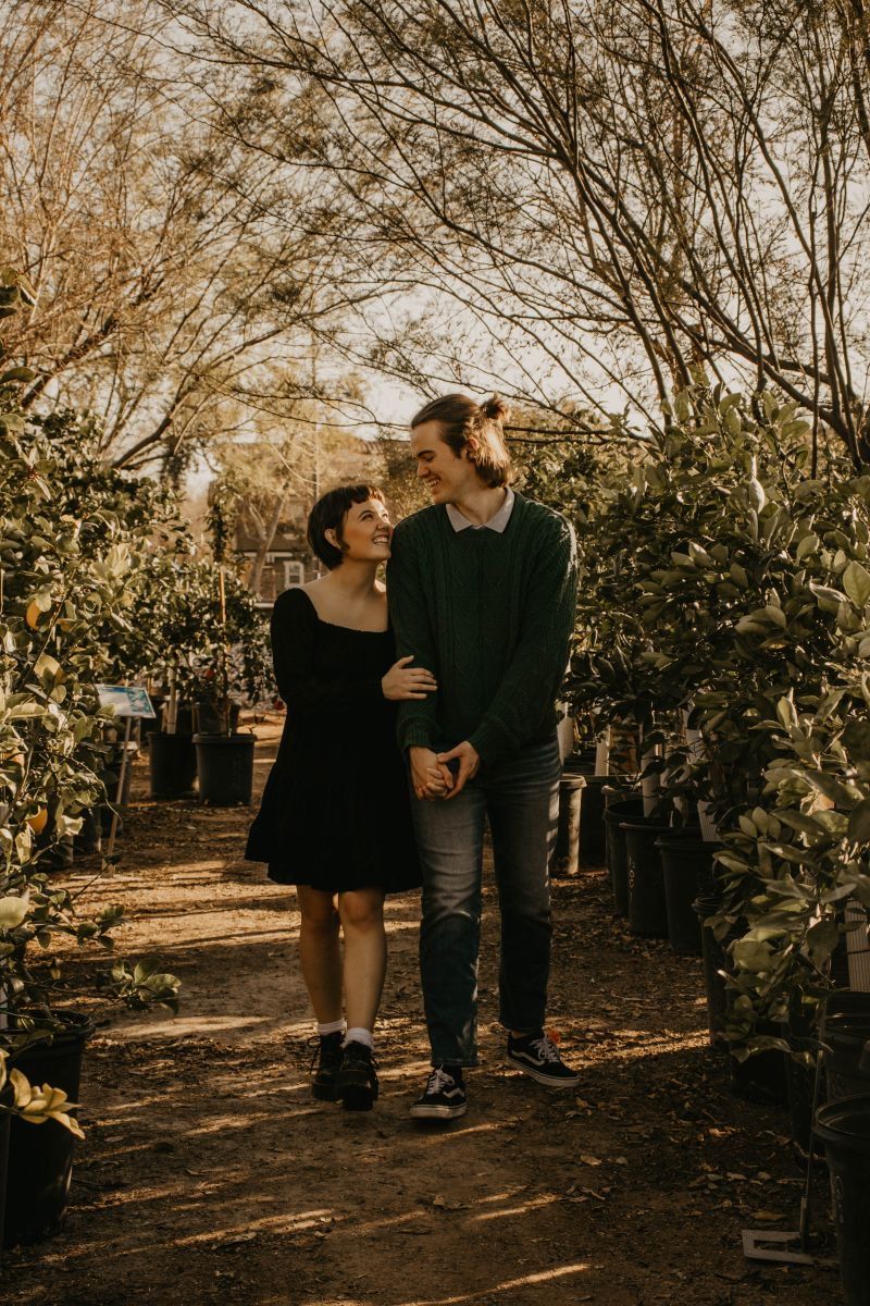 A couple walks hand-in-hand through a lush garden nursery, smiling at each other surrounded by potted plants.