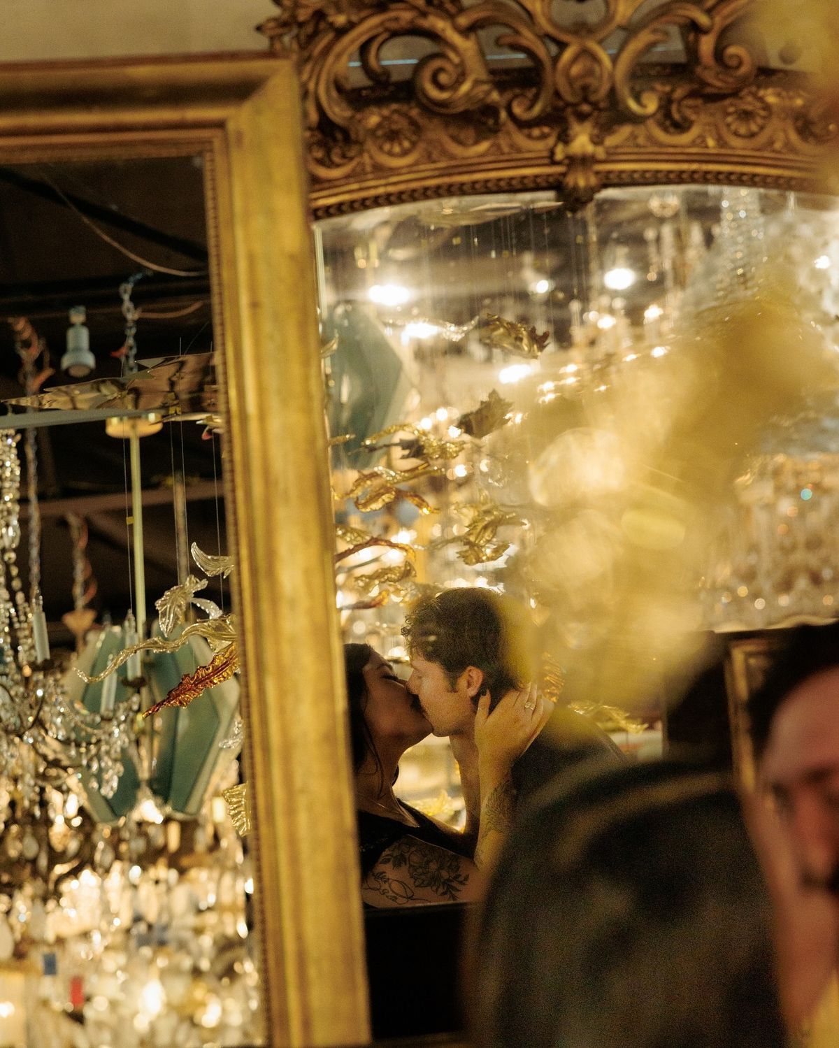 Couple kissing reflected in ornate gold mirror surrounded by sparkling chandeliers and glittering decorations.
