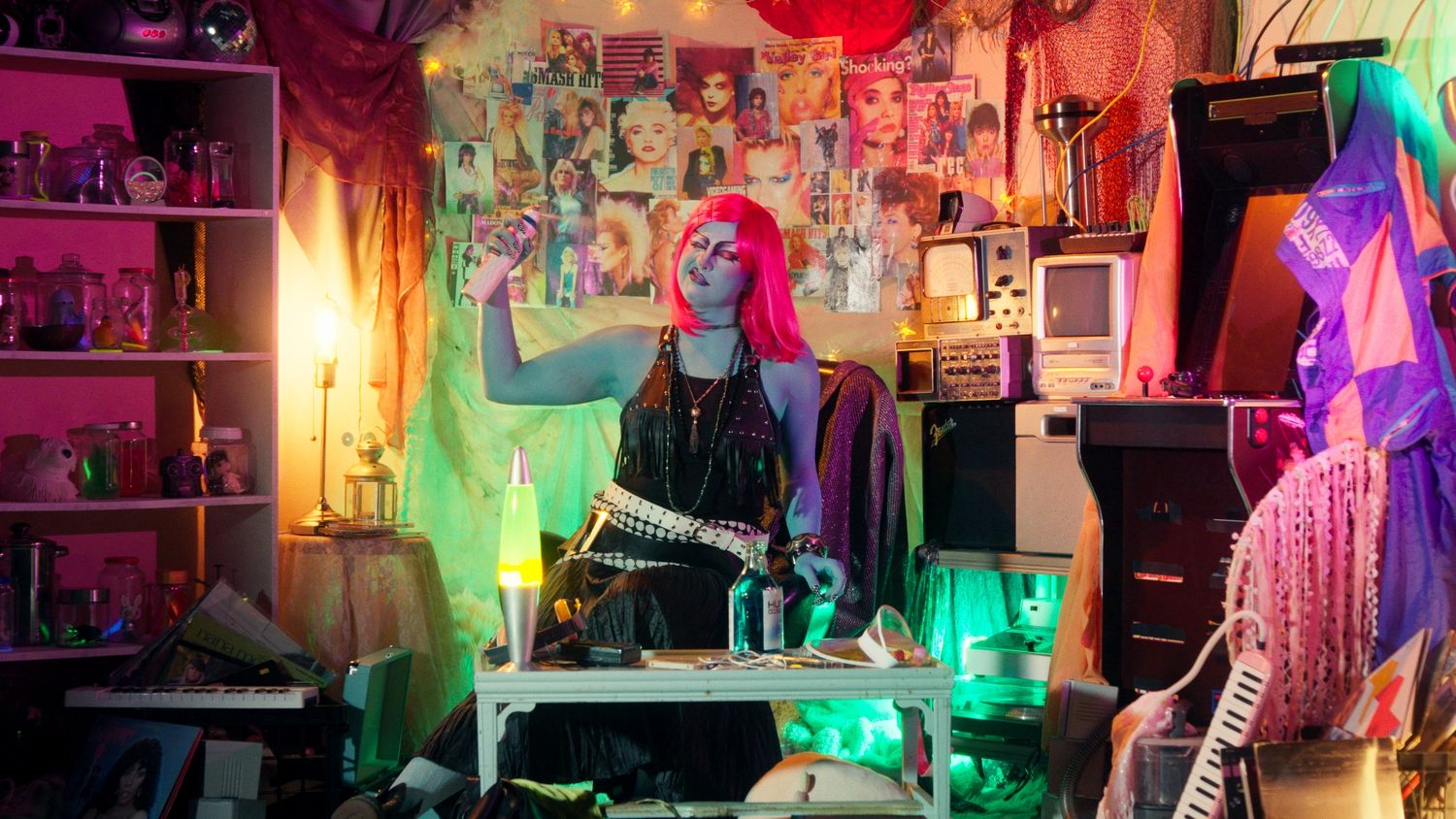 Person with pink hair and blue makeup poses in a colorful, eclectic room filled with posters and neon lights.