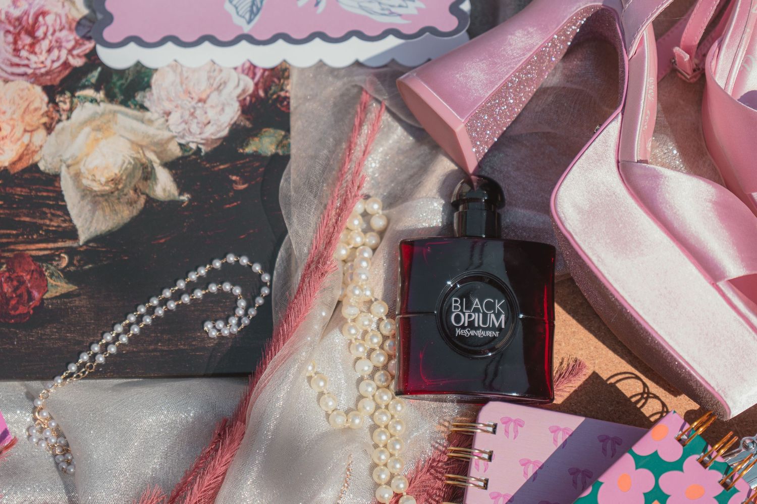 Flatlay of Black Opium perfume, pearl necklace, pink heels, floral book, and colorful accessories on white fabric.