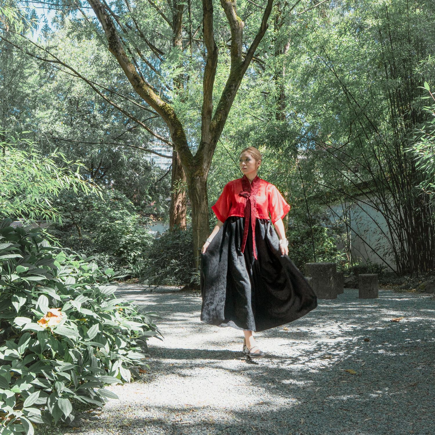 Woman in red top and black skirt walking on a garden path surrounded by lush green trees and white flowers.