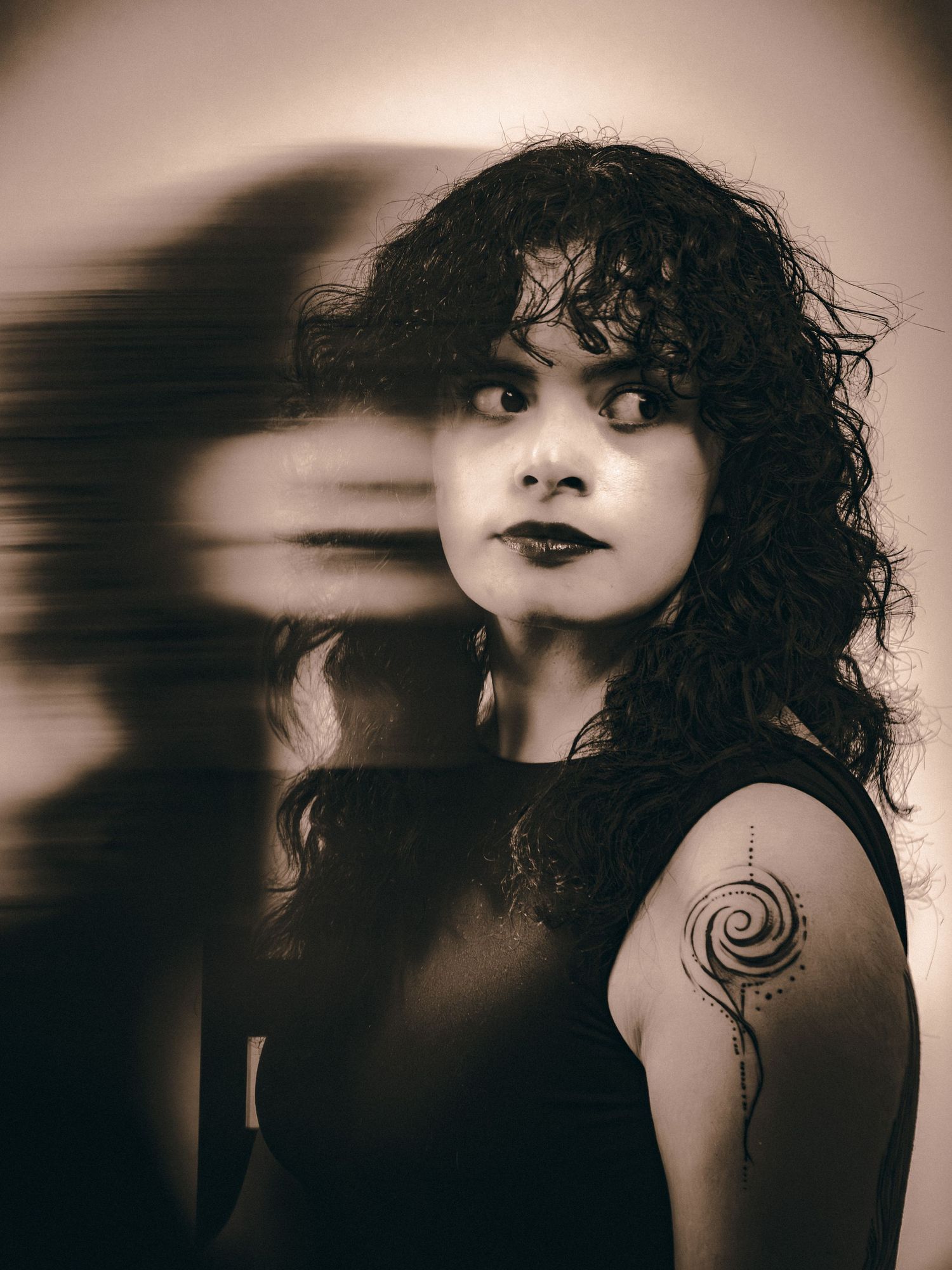 Artistic sepia double-exposure portrait of woman with curly hair and spiral shoulder tattoo in black.