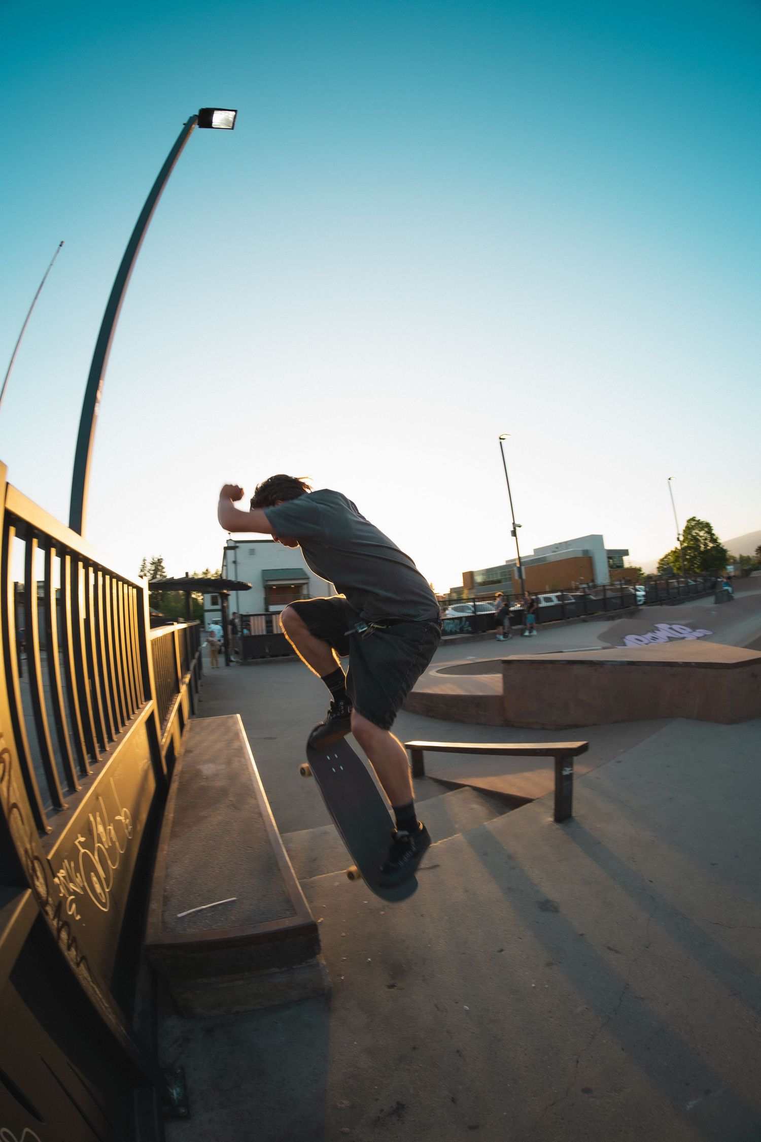 Skateboarder jumping near a railing at an outdoor skate park during warm sunset light.