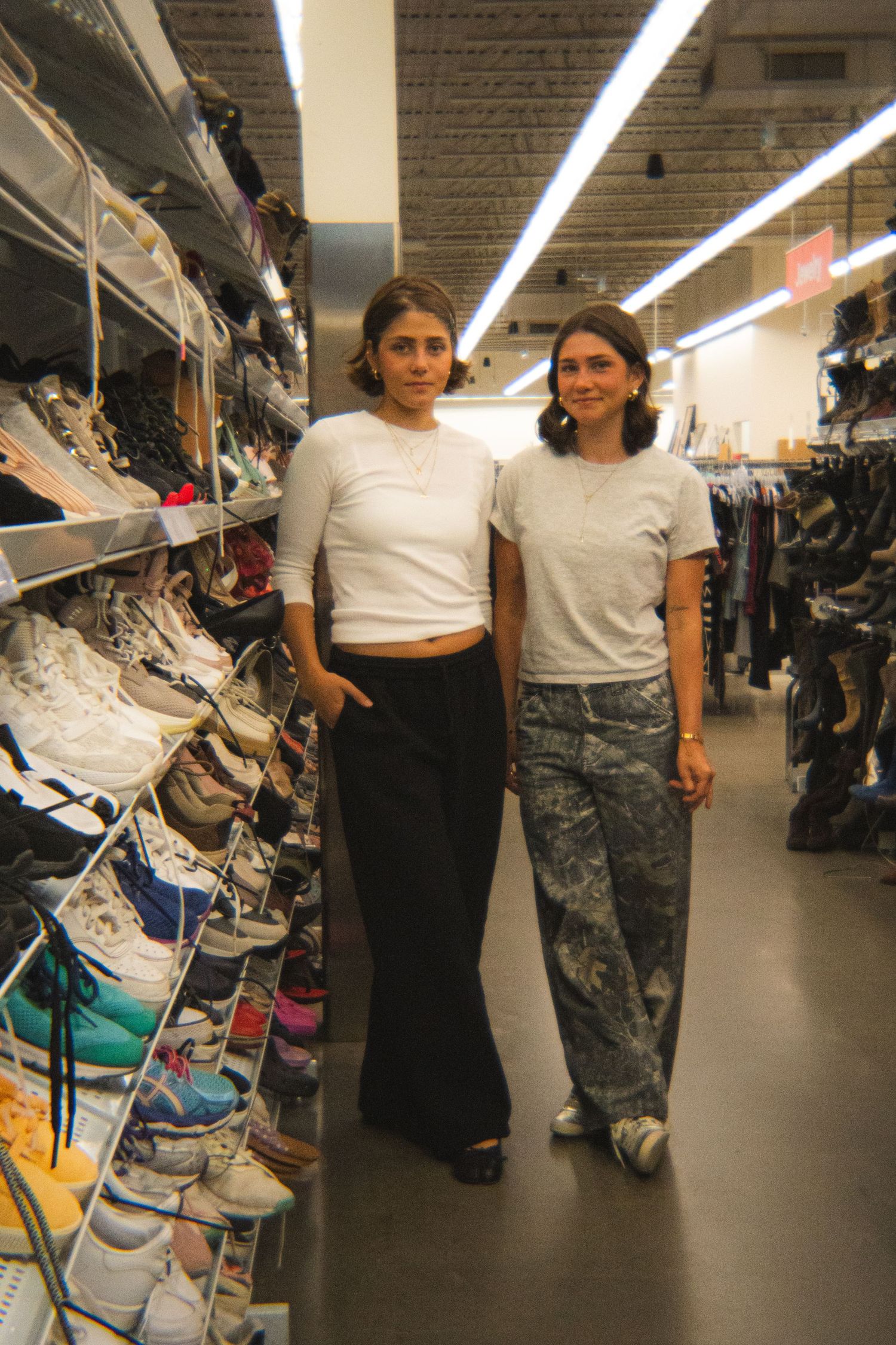 Two women in white tops pose together in thrift store shoe aisle with shelves of sneakers.