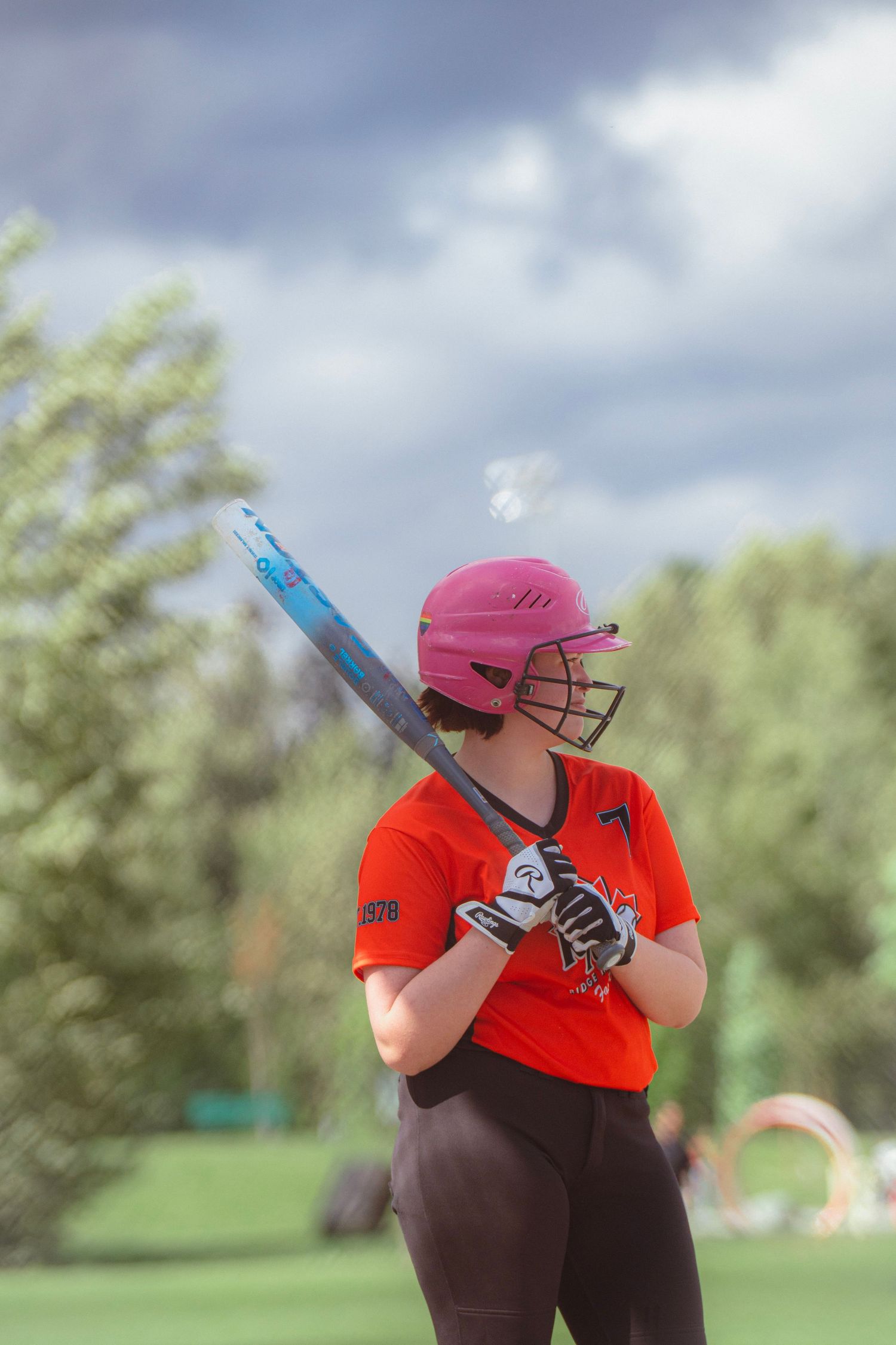 Female softball player in orange jersey and pink helmet holds blue bat ready to swing at outdoor field.