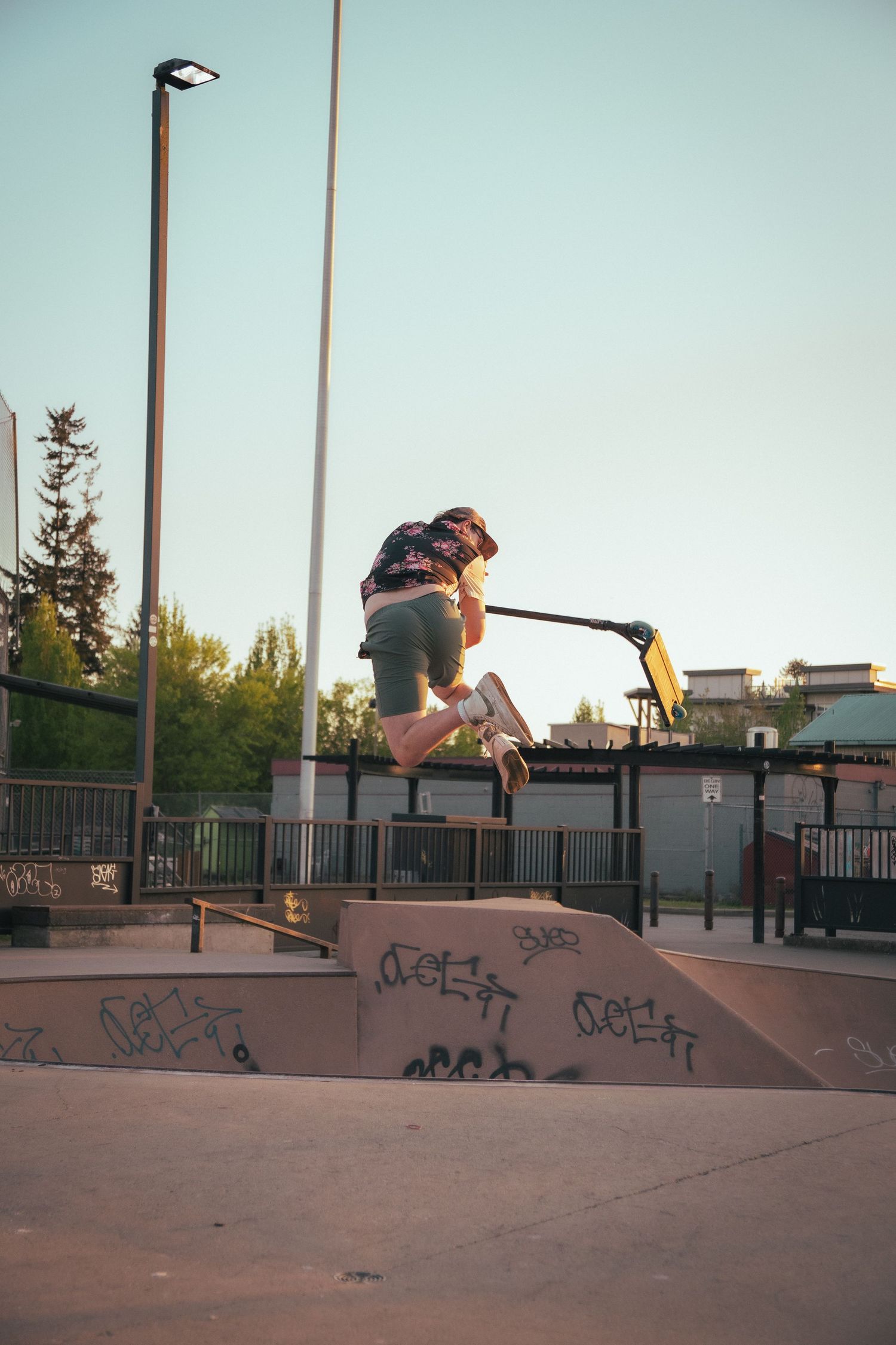 Rider performing a jump trick on a stunt scooter over a ramp at a skate park.