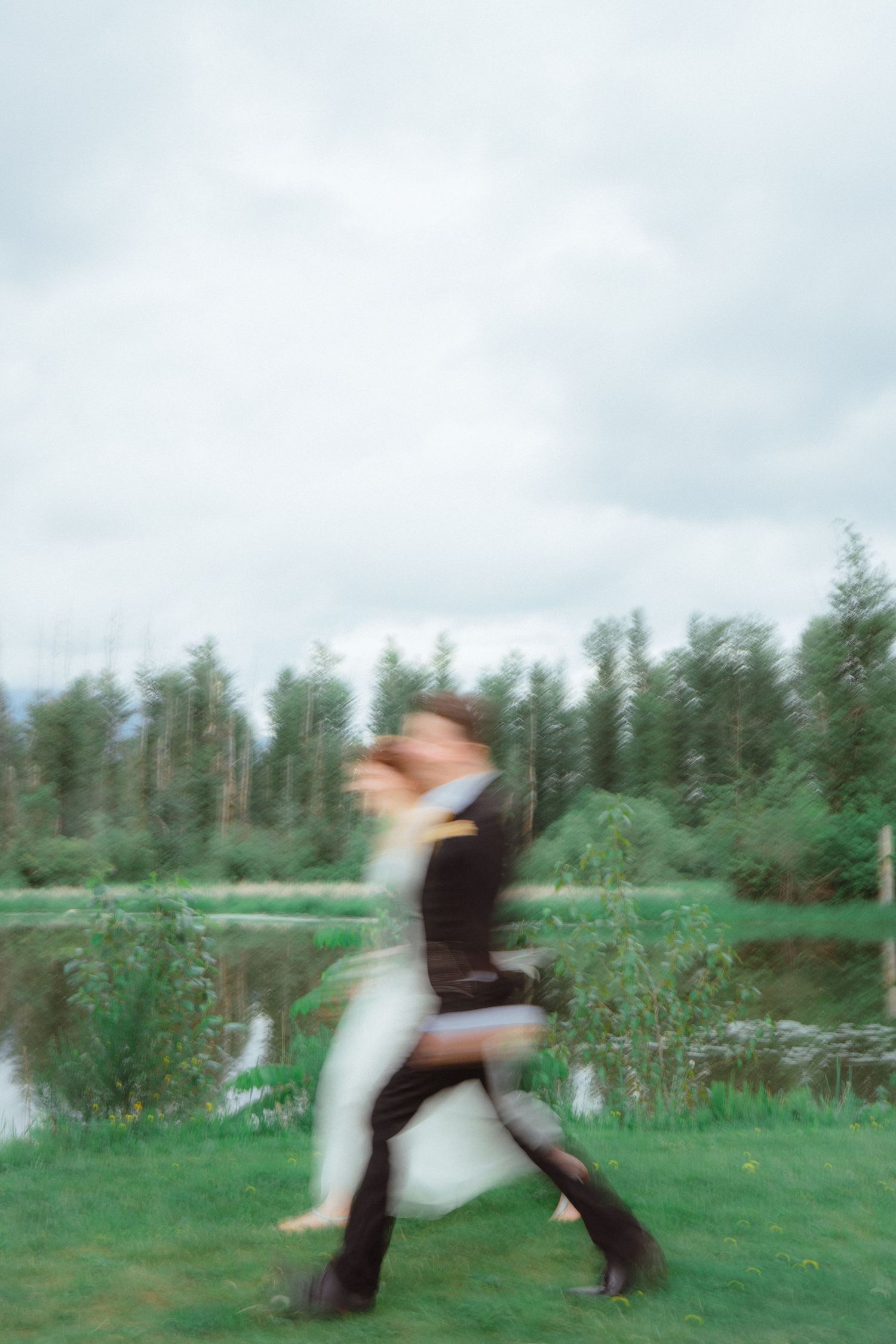 Motion blur photo of bride and groom dancing joyfully on green grass near tree-lined lake.
