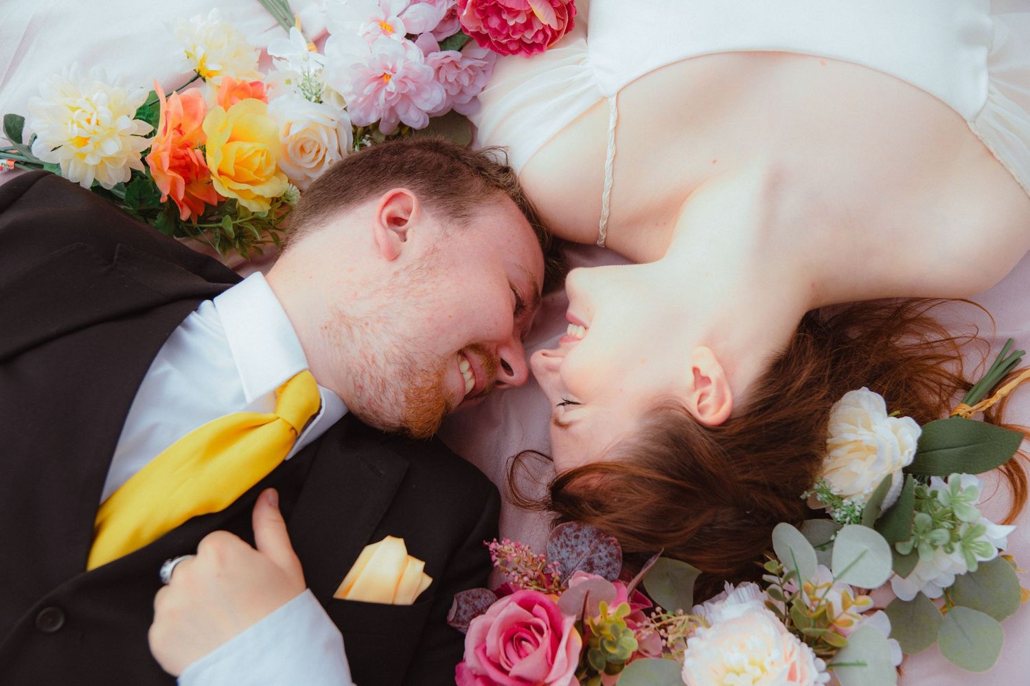 Bride and groom lying face to face smiling, surrounded by colorful wedding bouquets on pink fabric.