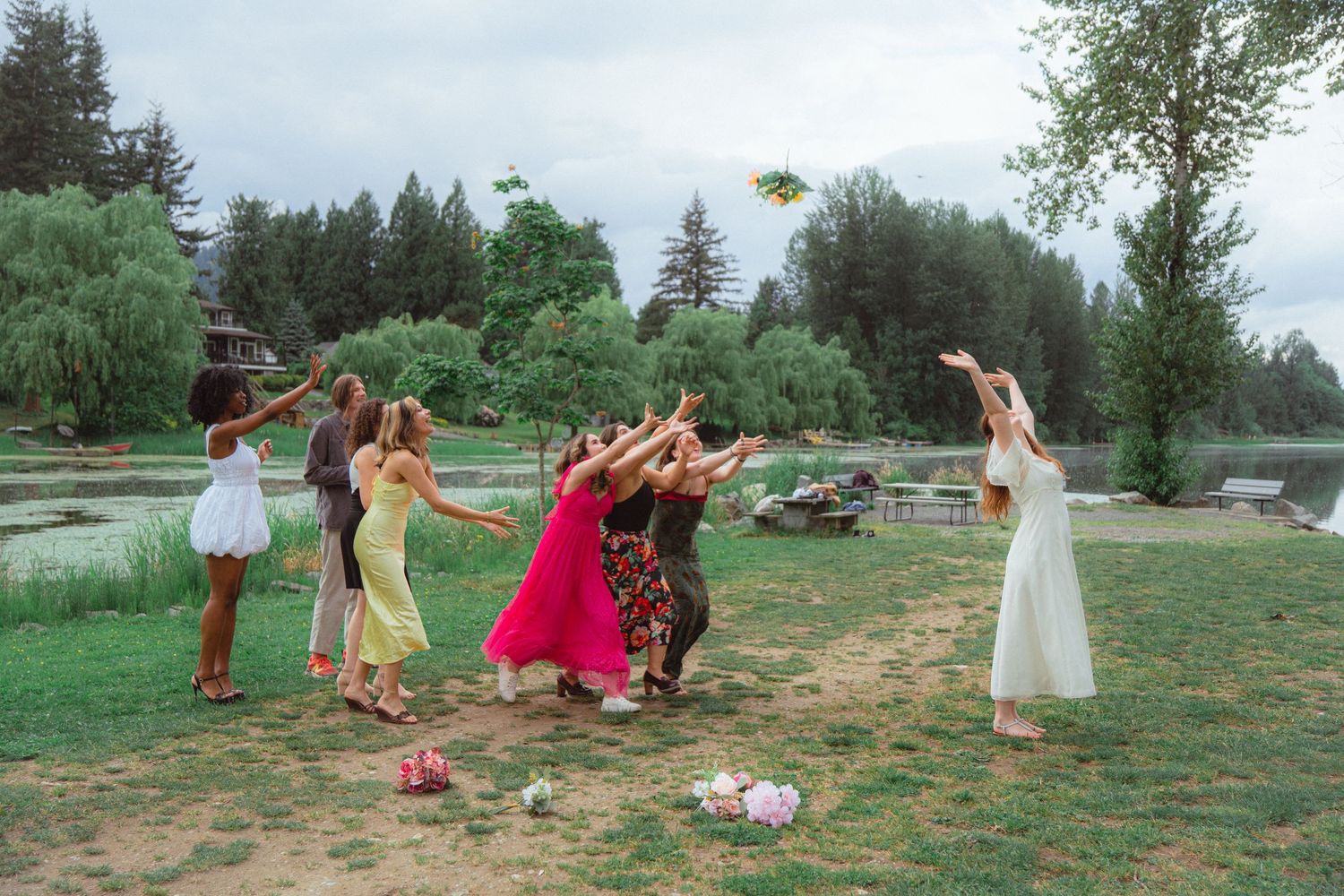 Wedding bouquet toss with bride throwing flowers to group of bridesmaids outdoors near lake.