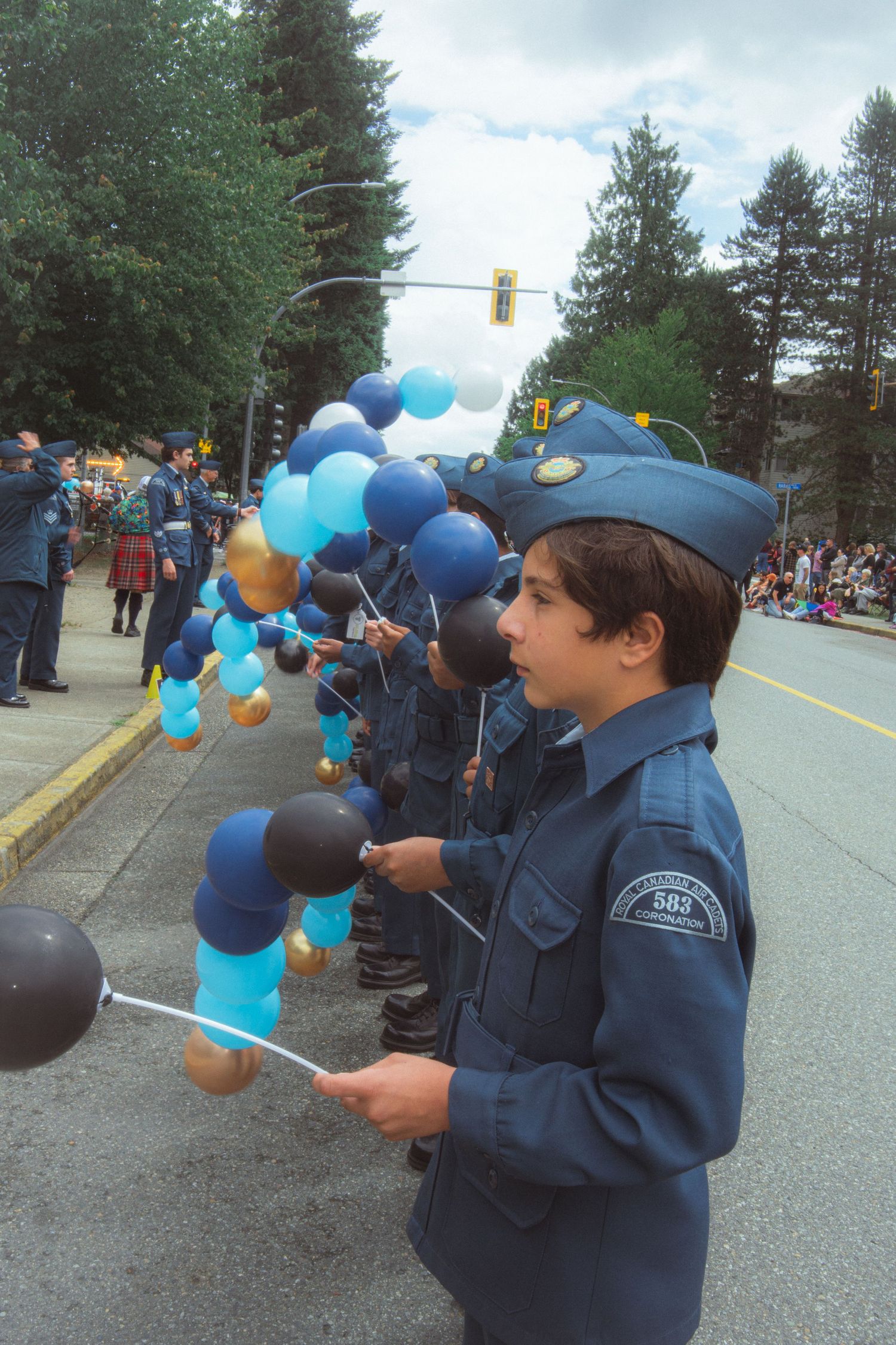 Young air cadet in blue uniform holds blue and gold balloons during a street parade.