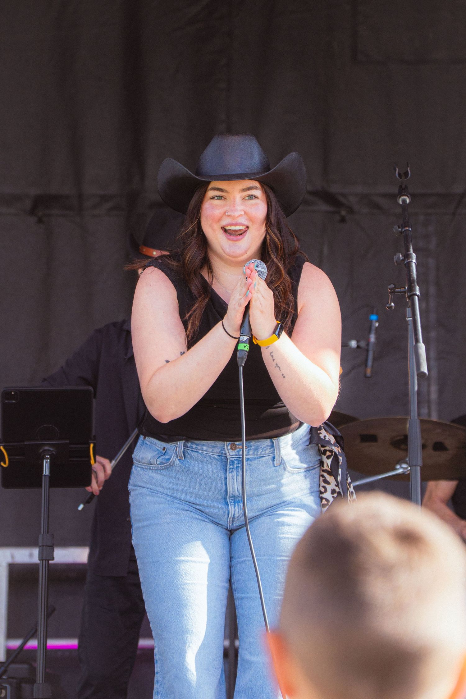 Smiling female country singer in black cowboy hat clasps microphone on sunny outdoor festival stage.