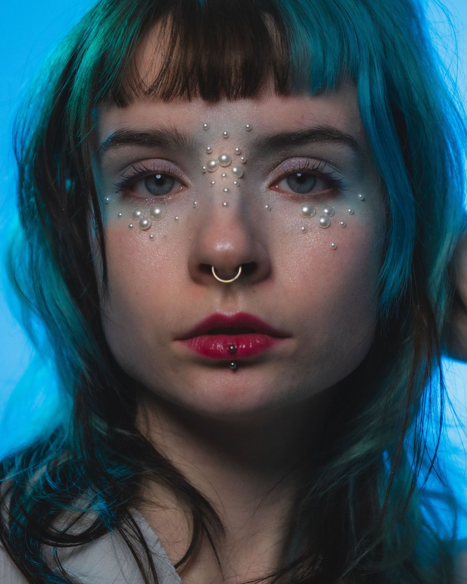Portrait of woman with teal hair, nose ring, and pearl face gems on blue background.