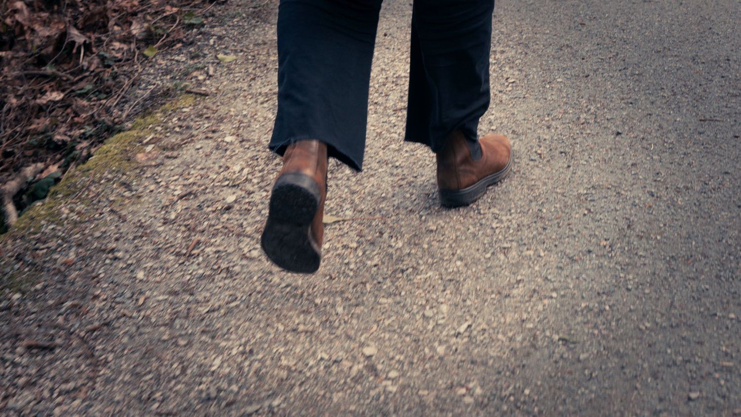 Close-up of a person walking on a gravel path wearing dark pants and brown leather shoes outdoors.