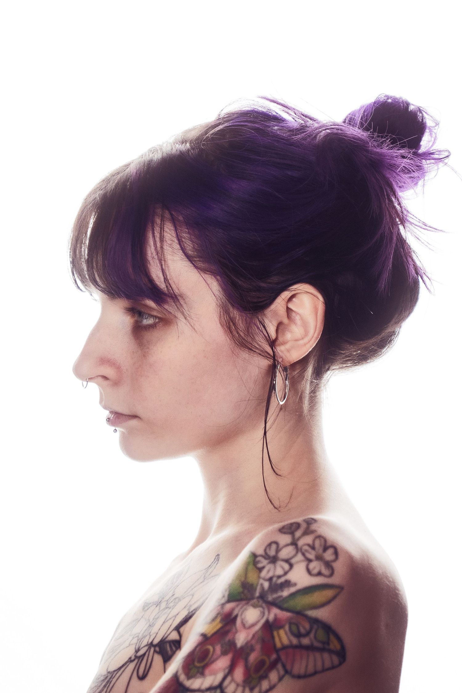 Profile view of woman with dark purple updo hair and colorful shoulder tattoos against white background.
