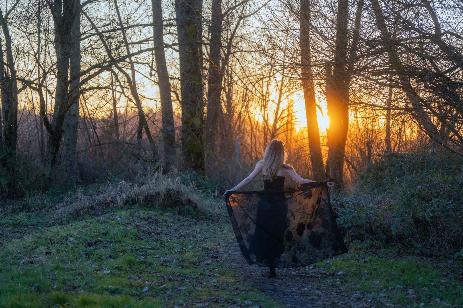 Woman in black dress spreading her shawl wide in a forest clearing at golden sunset.