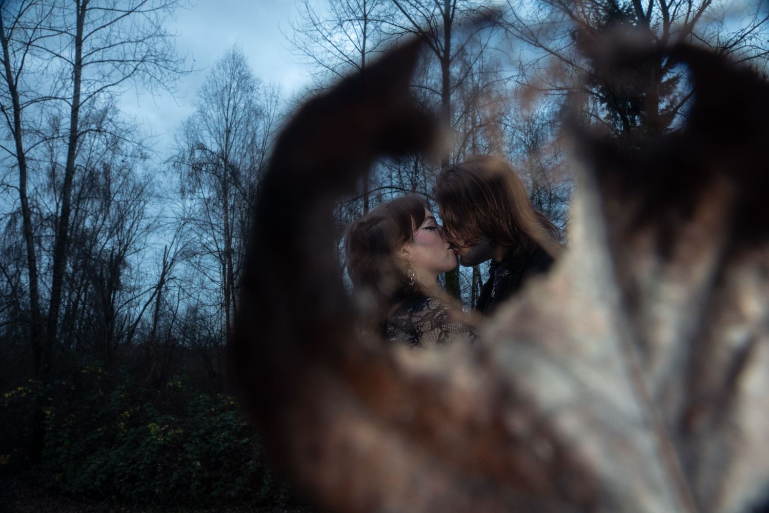 A couple shares a romantic kiss in a forest, photographed through a blurred autumn leaf in the foreground.