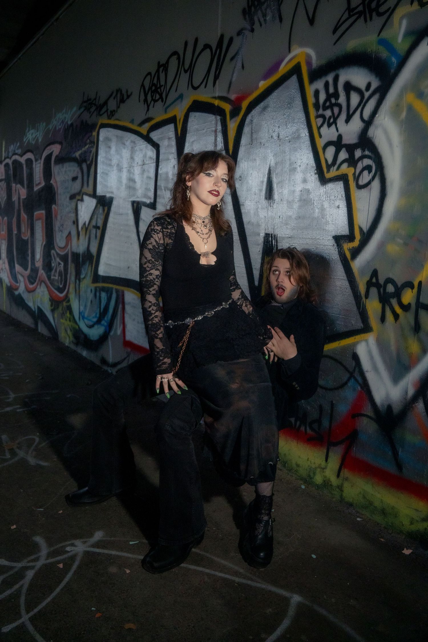 Gothic woman in black lace top poses with crouching man against vibrant graffiti wall.