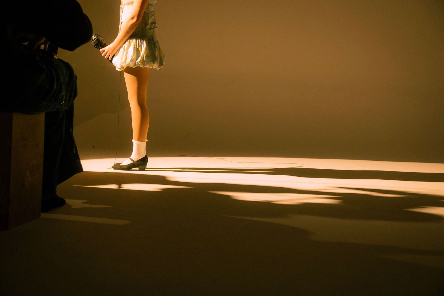 A dancer in a white skirt and black shoes stands in a warm spotlight on a golden-lit stage floor.