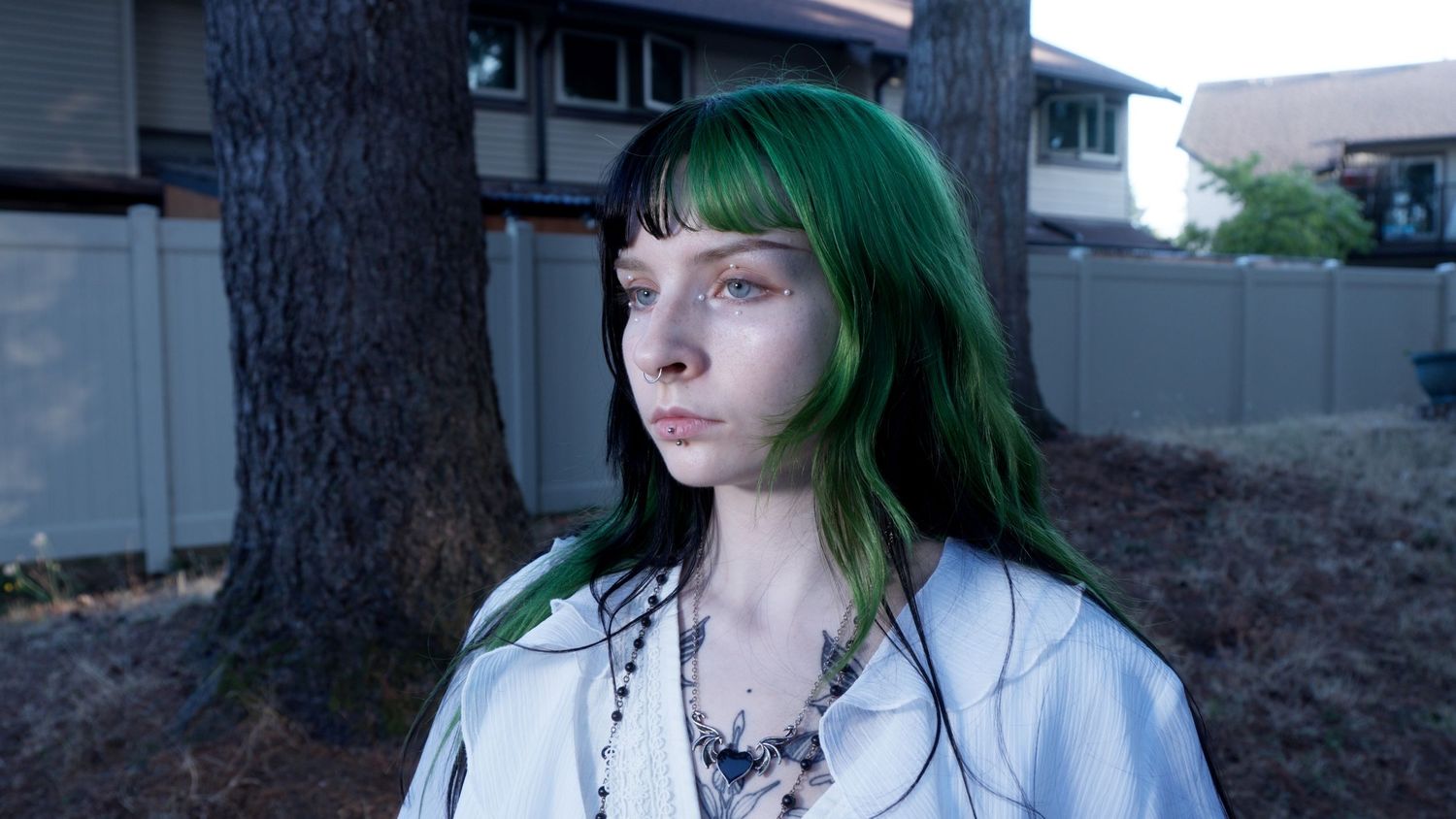 Young woman with green hair and necklaces in white shirt stands outdoors near tree.