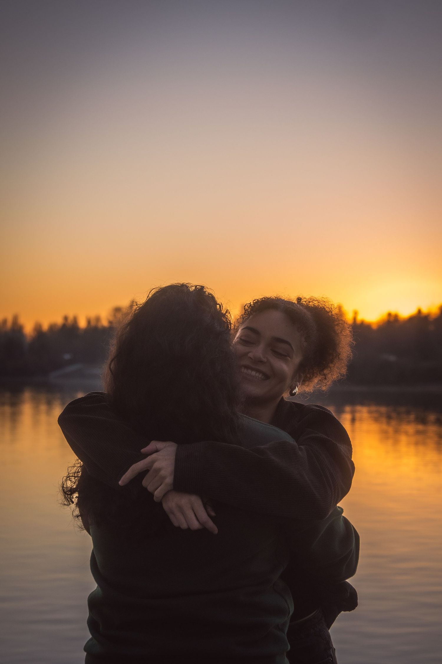 Two women hugging joyfully by a lake during a golden sunset.