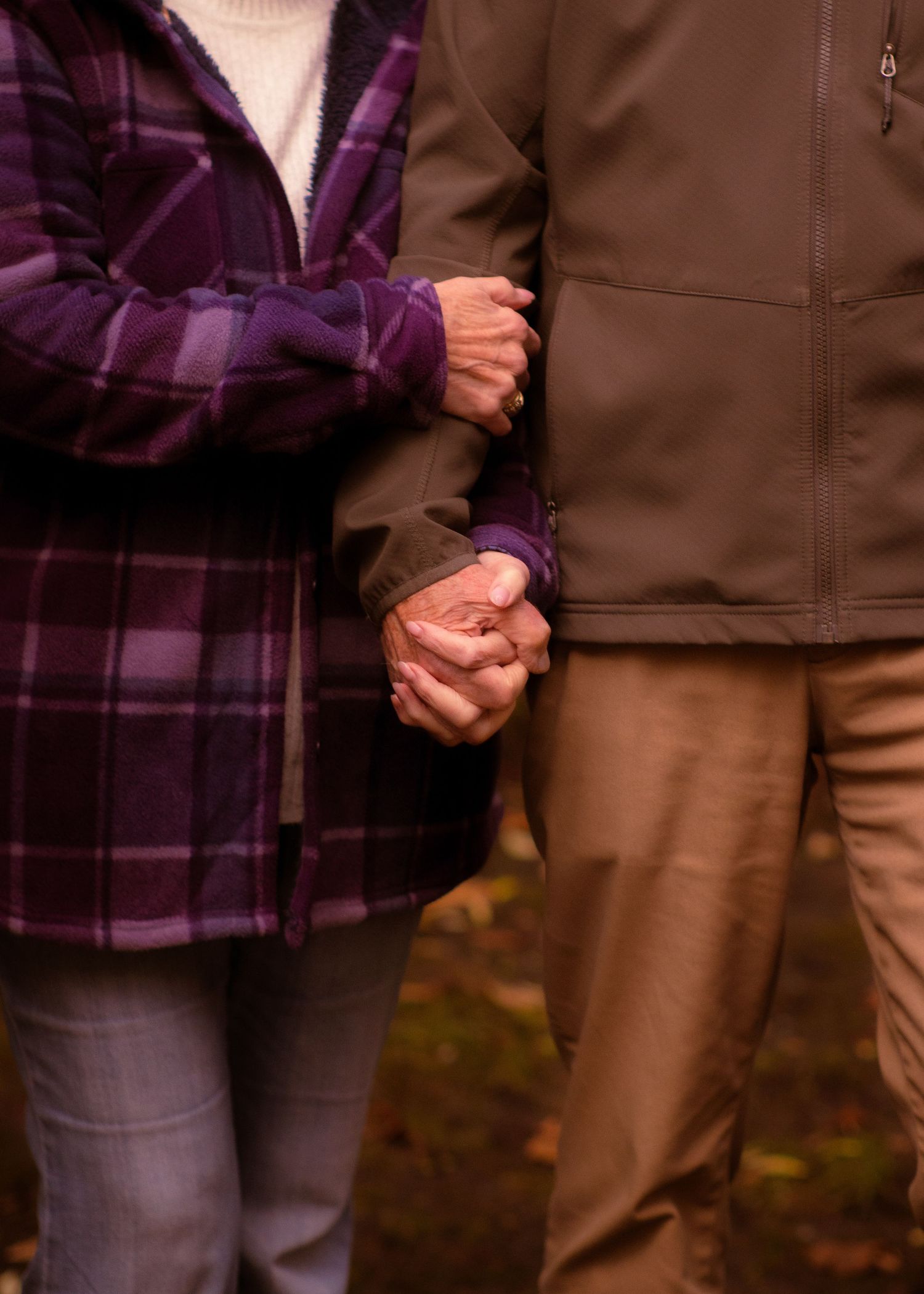 Senior couple holding hands outdoors in autumn, wearing purple plaid and brown jacket, showing enduring romance.