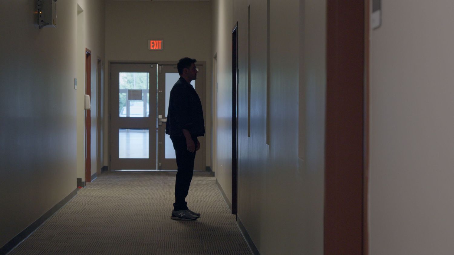 Silhouetted person standing in a dimly lit office hallway with an illuminated exit sign above a door.