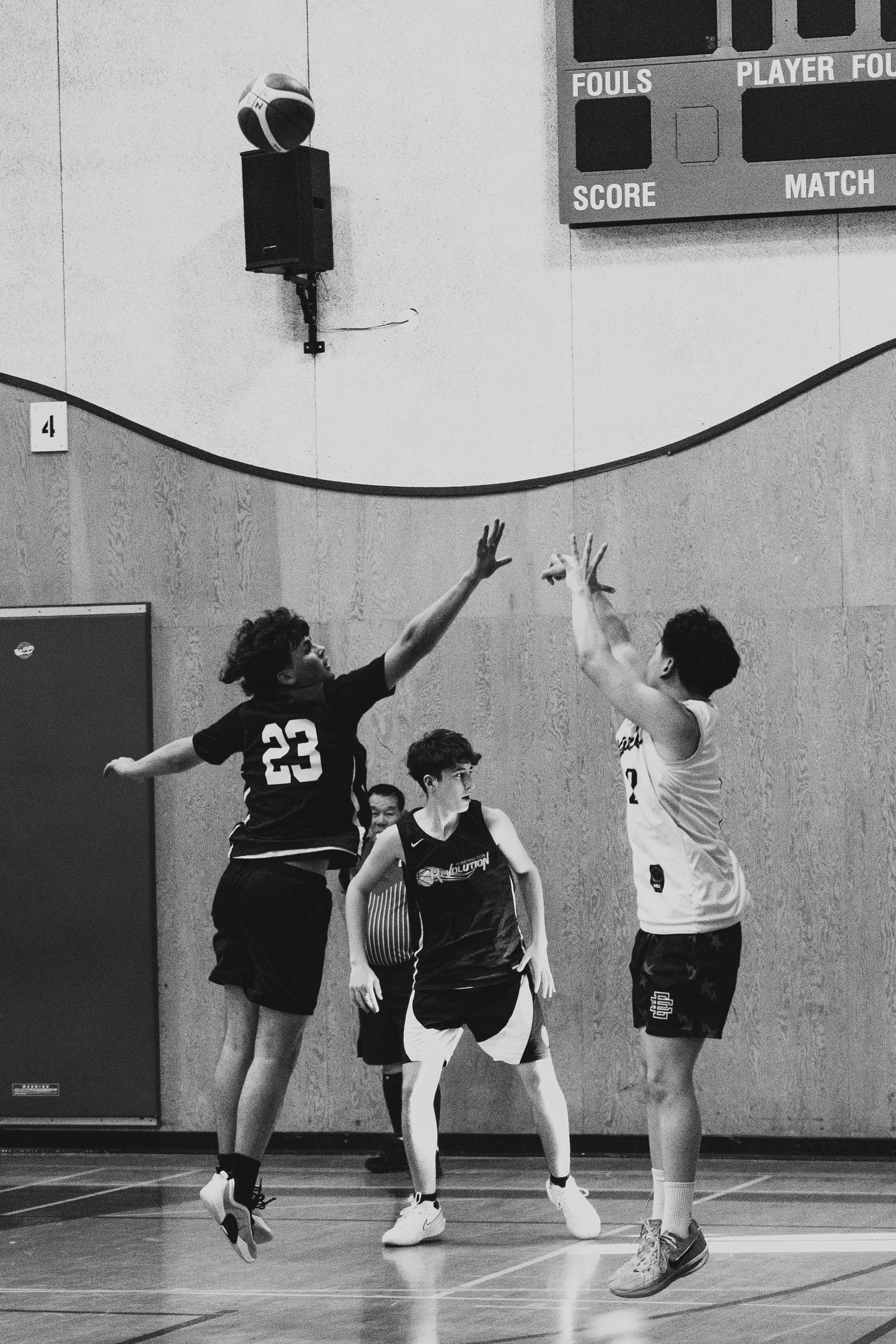 Player #23 leaps to contest a jump shot during indoor basketball game near scoreboard.