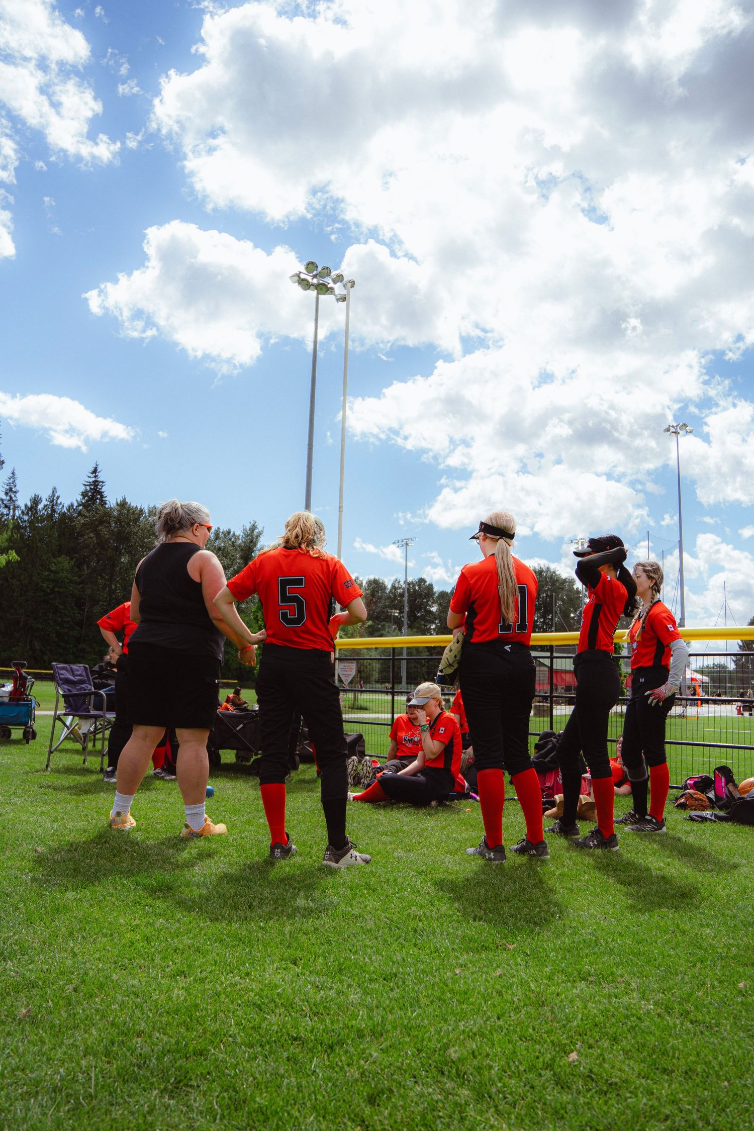 Female softball team in orange uniforms having discussion on green field under cloudy blue sky.