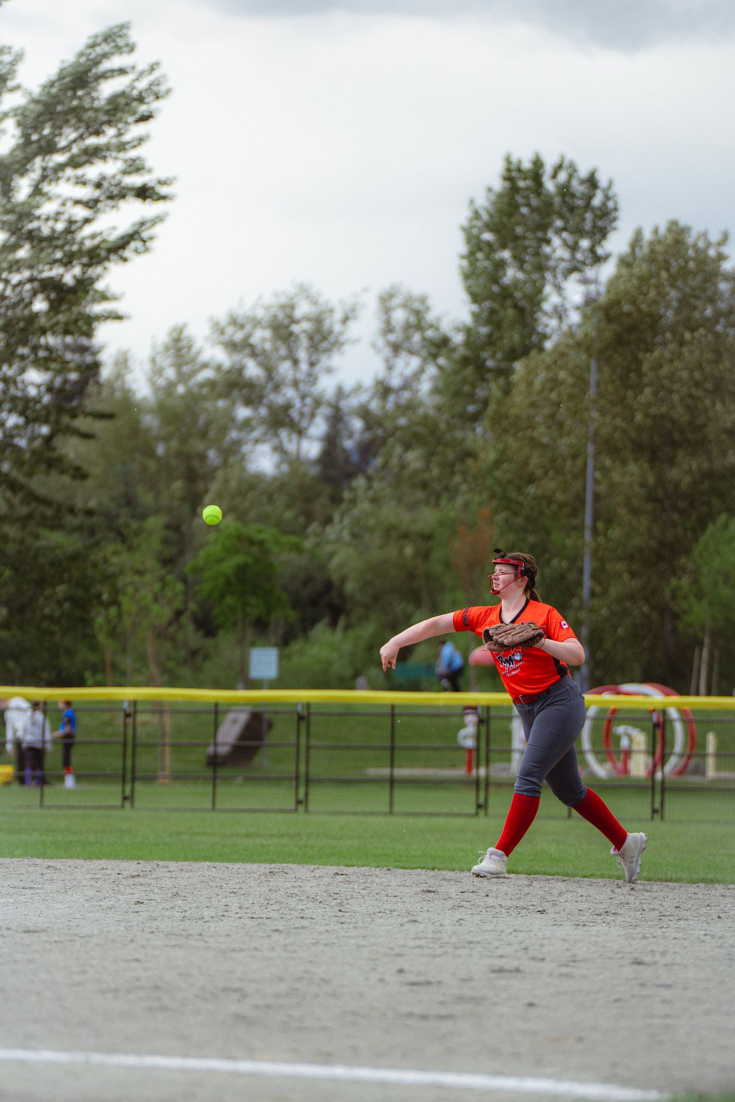 Female softball player in orange uniform throws the ball across the field during an outdoor game.