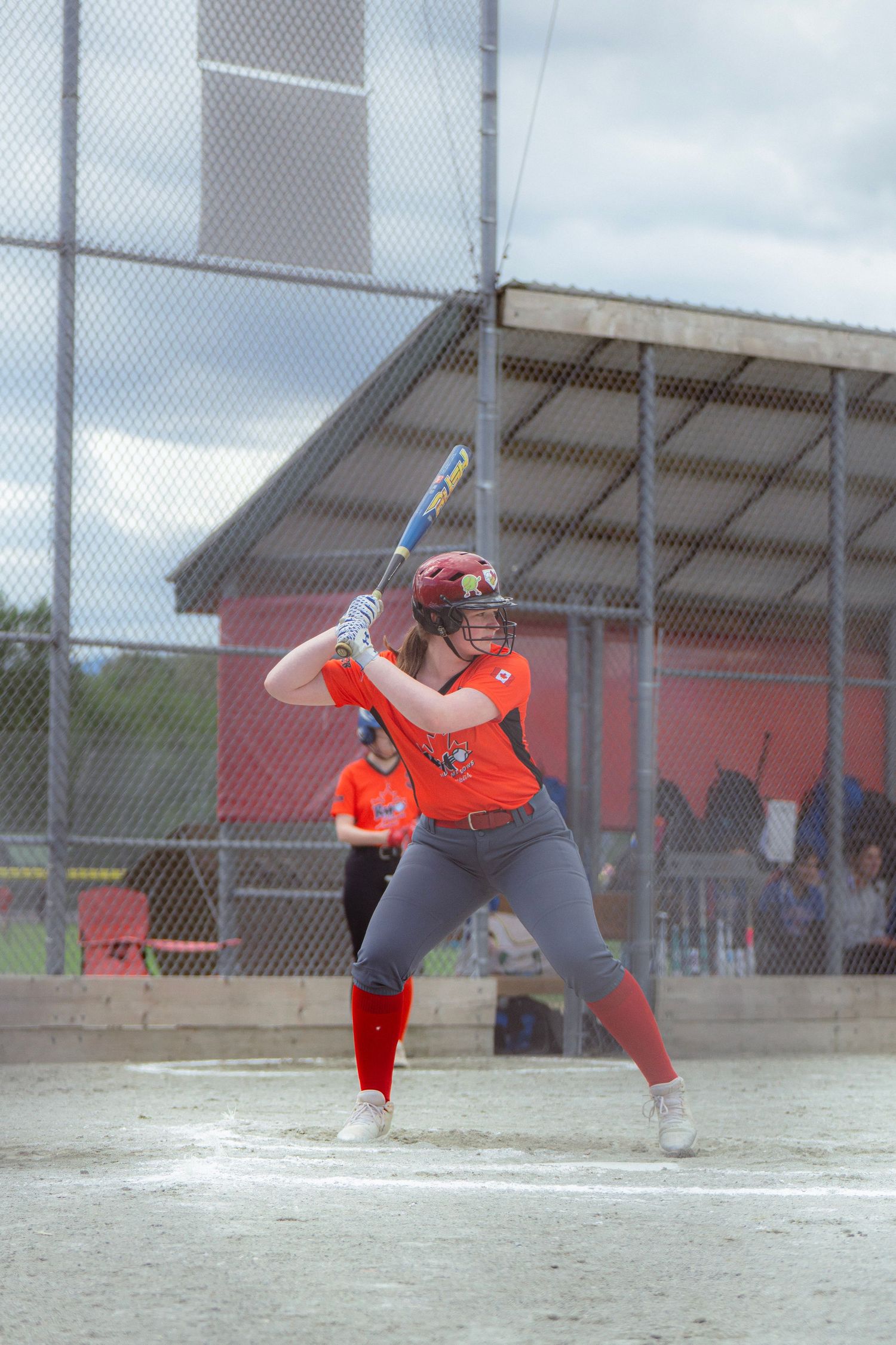 Female softball batter in orange uniform raises bat in ready position during outdoor game.
