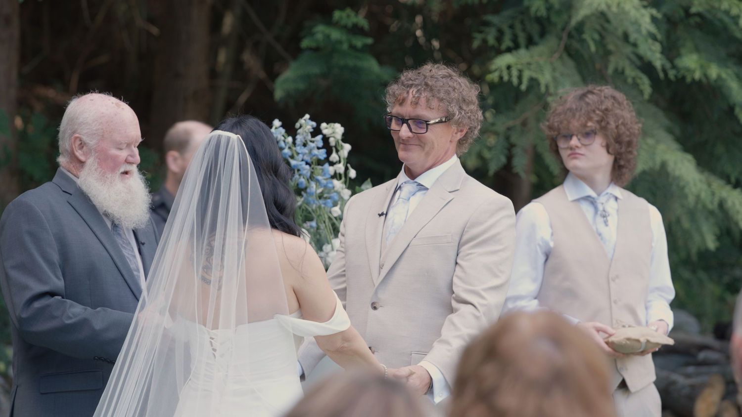 Groom in beige suit exchanges vows with bride in white gown at outdoor wedding ceremony.