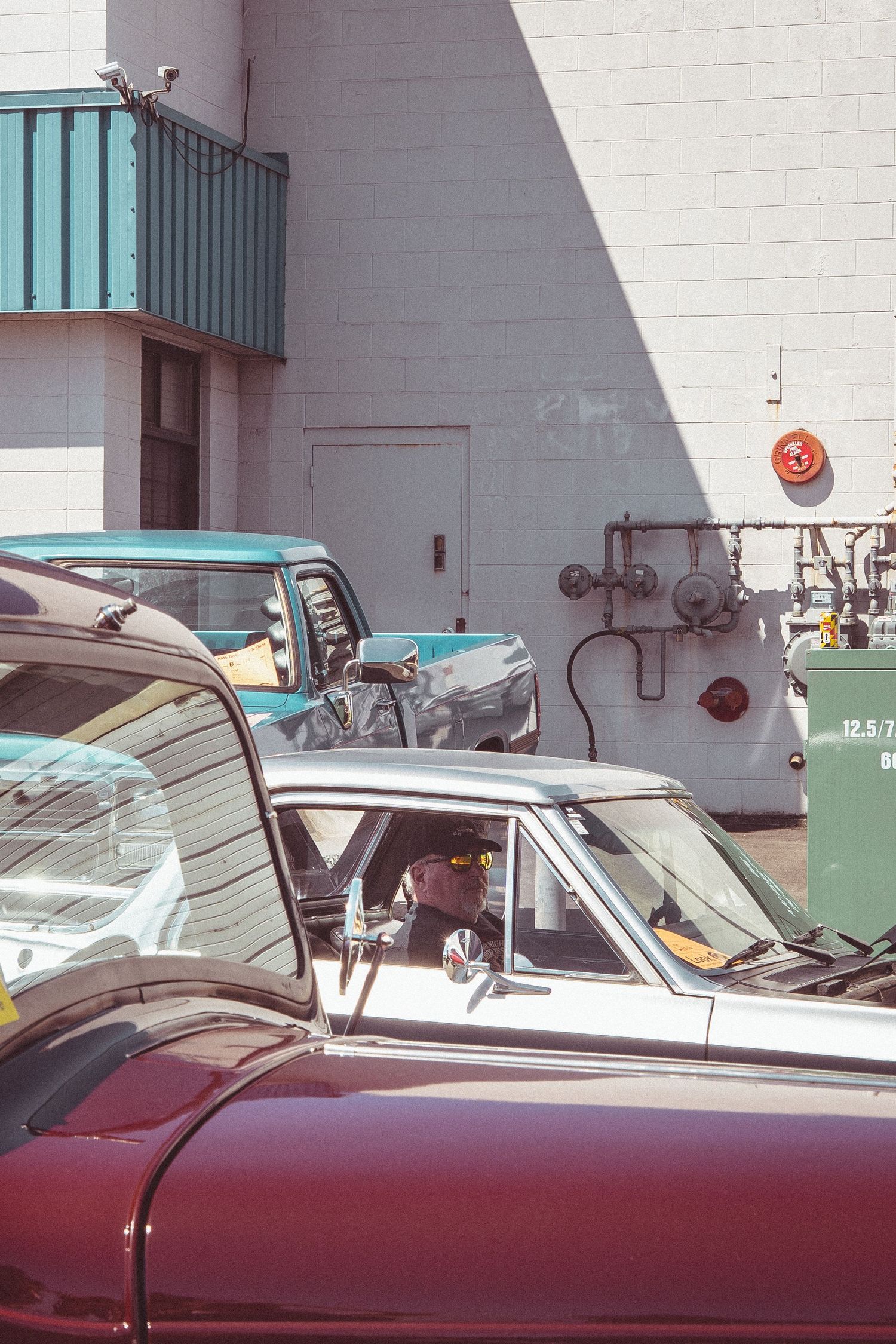 Man wearing sunglasses sitting in a classic convertible car at a vintage auto show near a building.