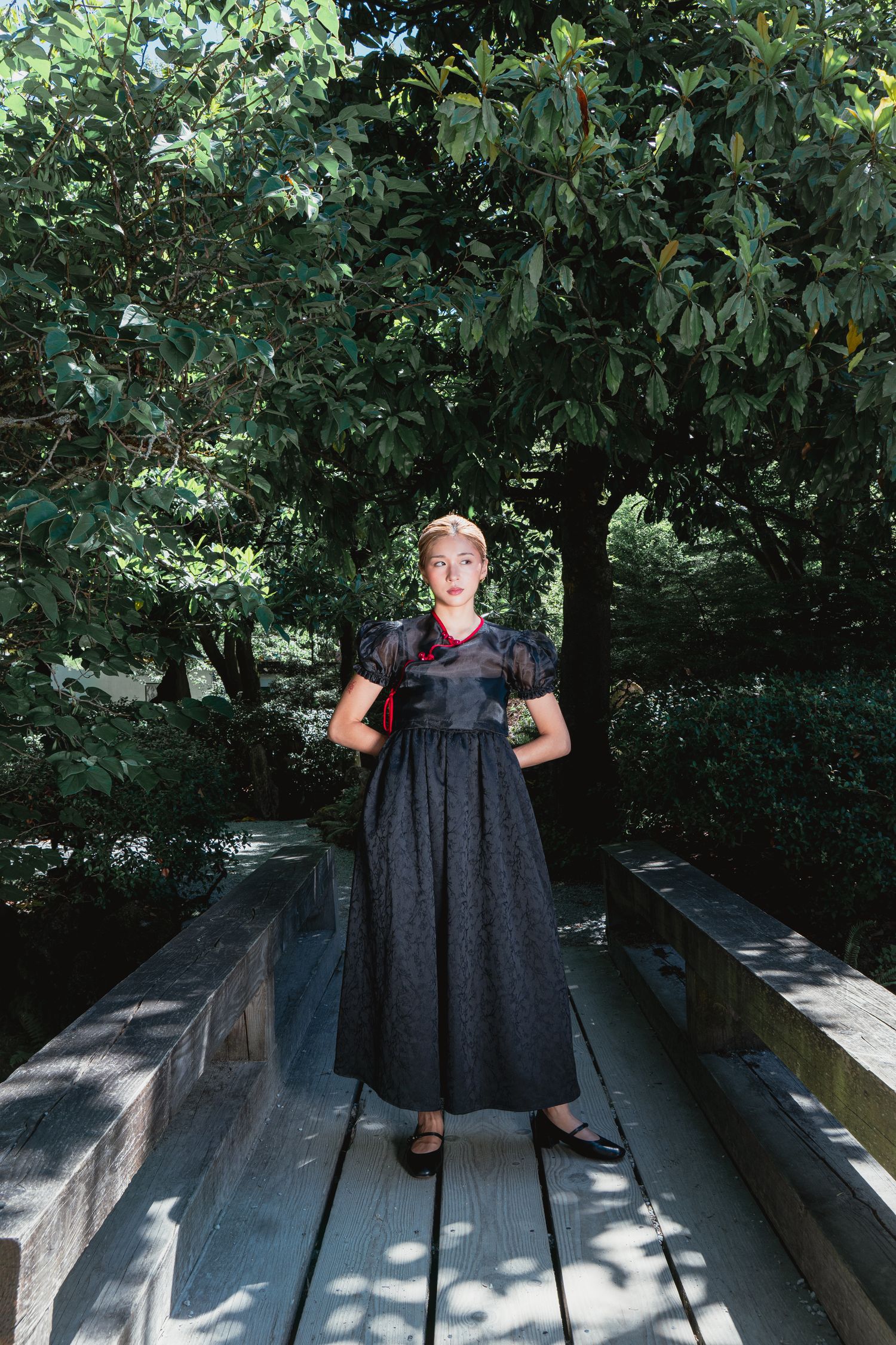Woman in dark floral midi dress standing on wooden bridge surrounded by lush green trees in a garden.