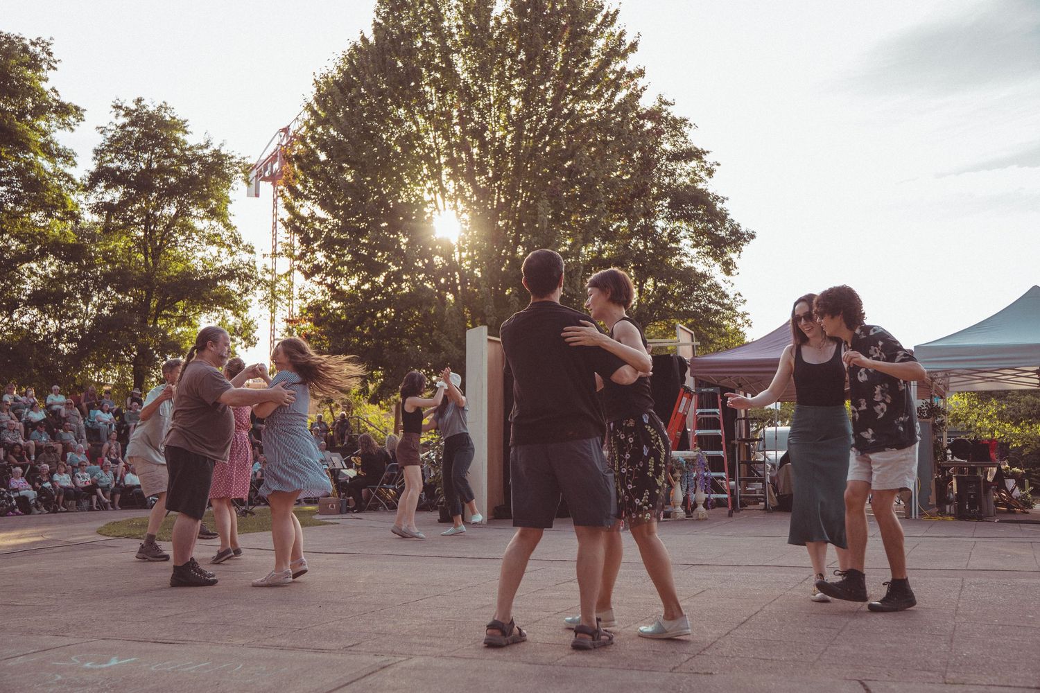 Couples dancing outdoors at a summer festival with sunlight filtering through trees at golden hour.