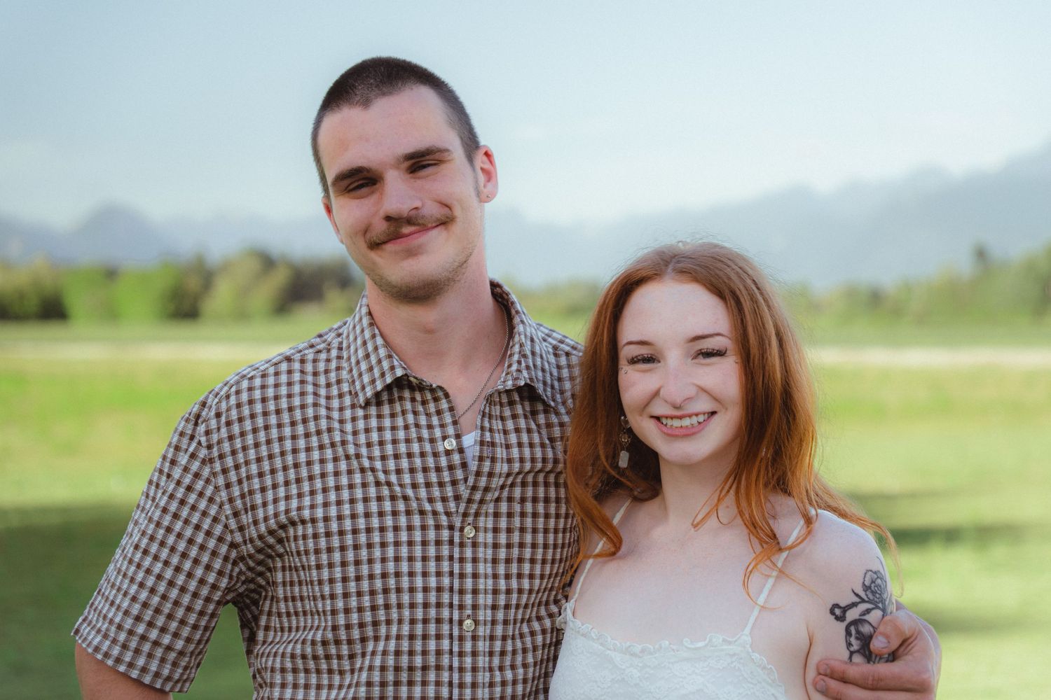A smiling young couple poses outdoors with green fields and mountains in the background on a sunny day.