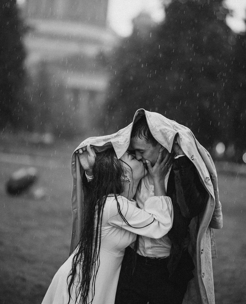 Couple sharing a passionate kiss in the rain while sheltering under a coat in black and white photo.