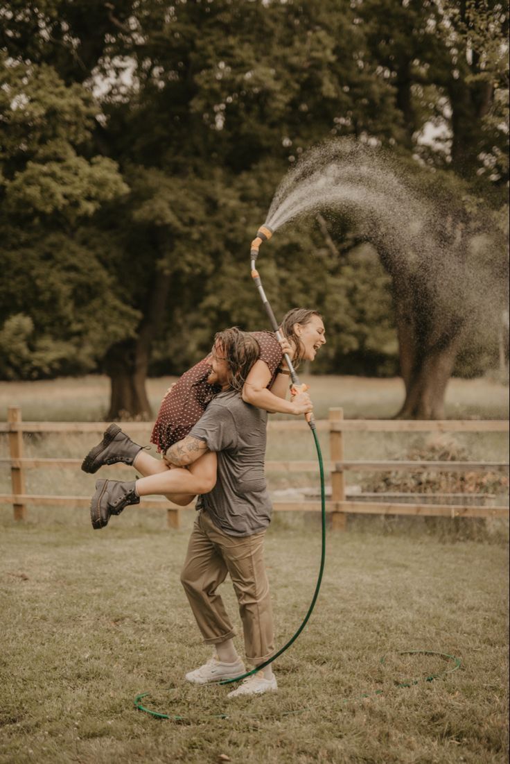 Playful couple with garden hose, woman held aloft spraying water in lush green field.