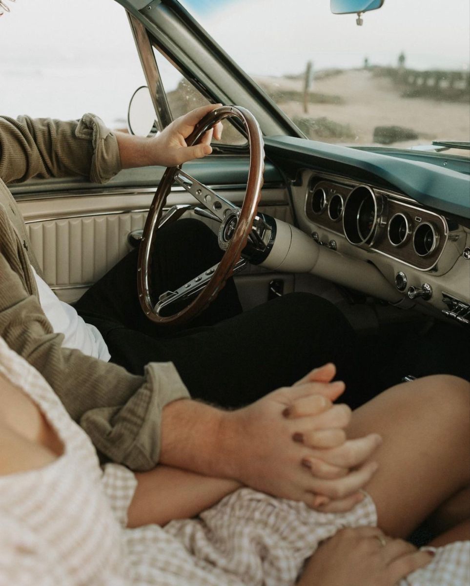Couple holding hands in a vintage classic car near the beach, with one person gripping the steering wheel.
