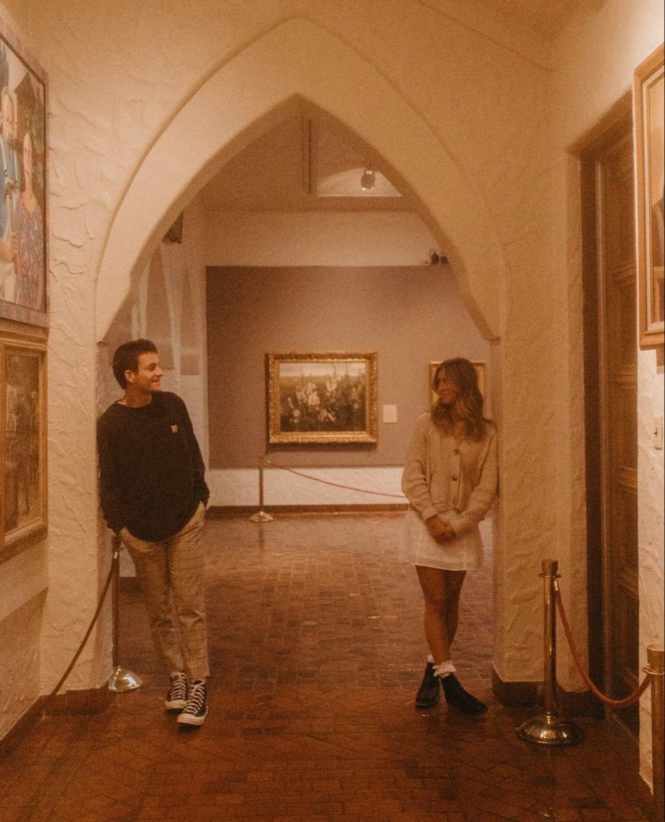 Two visitors conversing in a museum hallway with arched doorway, artwork on walls, and rope barriers.