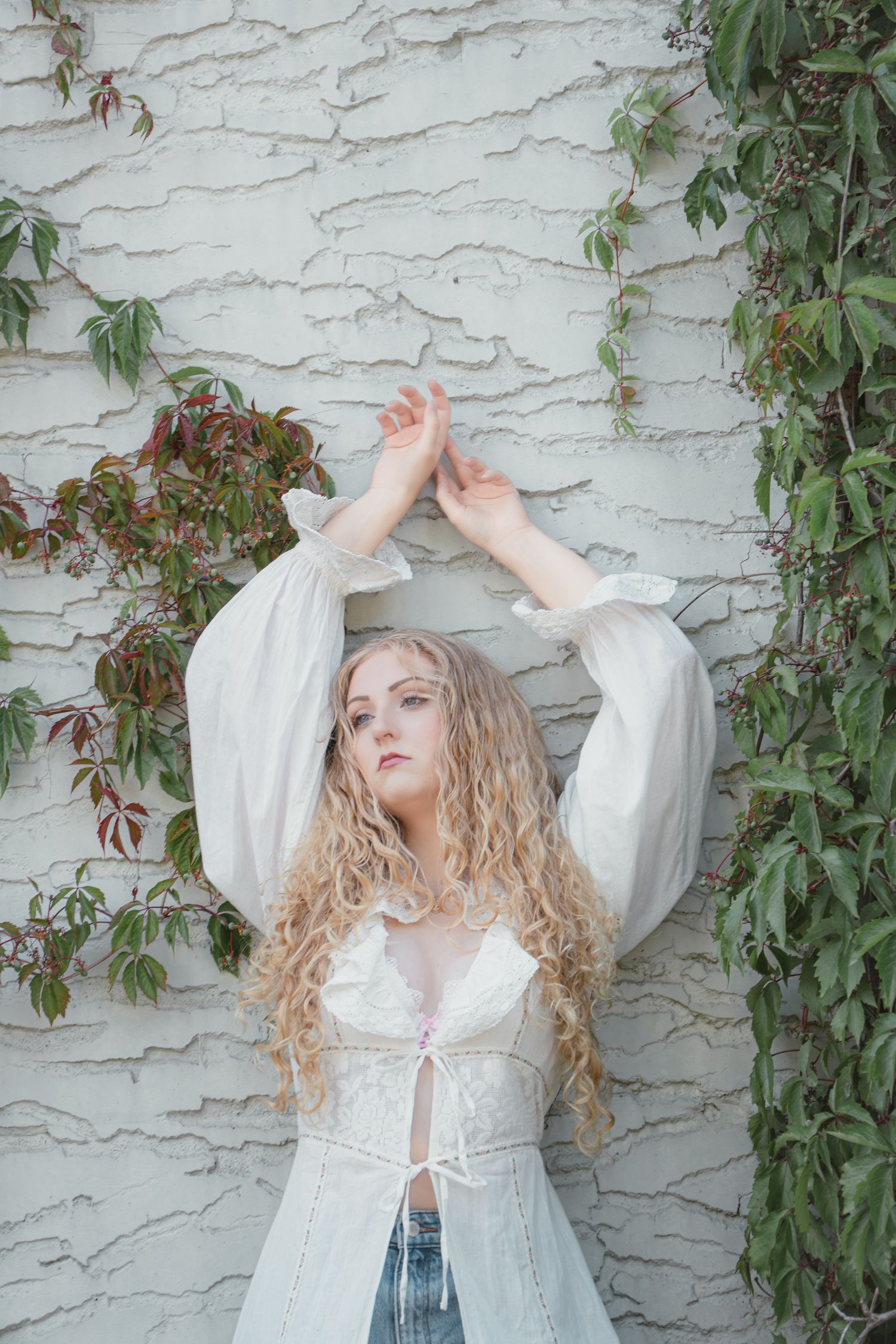 Young woman with curly blonde hair in white lace top raises arms against ivy-covered white textured wall.