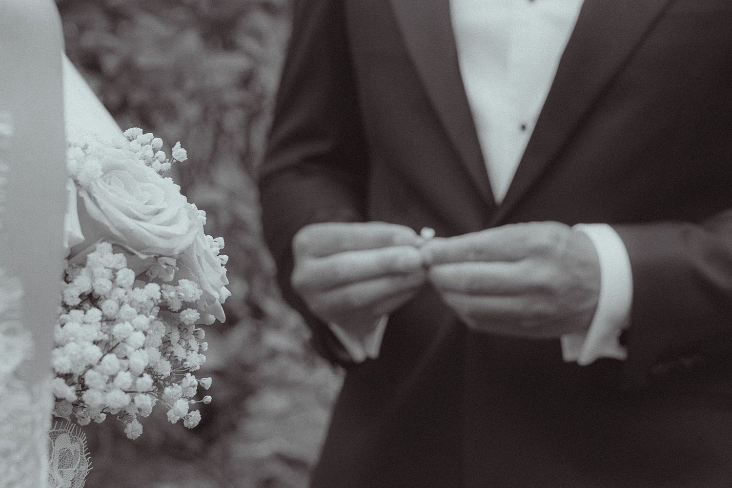 Black and white photo of groom adjusting cufflinks beside bride holding white rose bouquet at wedding ceremony.