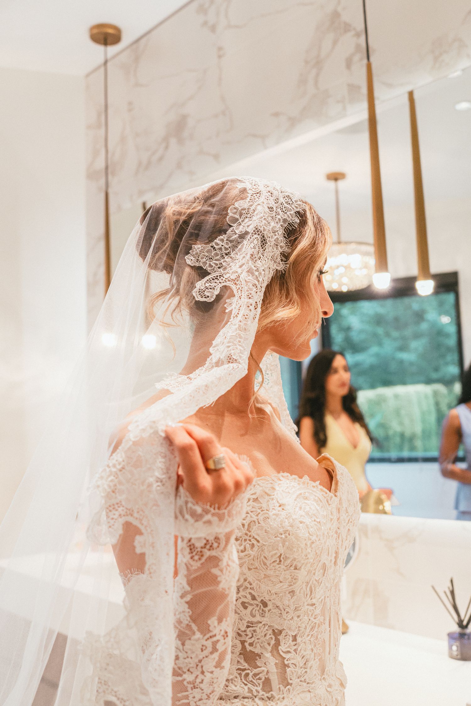 Bride in a lace wedding gown and veil gazing over her shoulder in a luxurious marble bathroom.