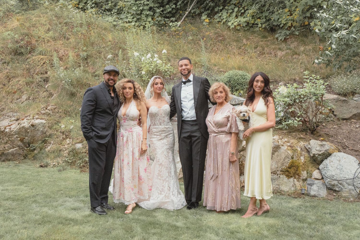 Wedding party posing outdoors on green lawn, bride in lace gown, groom in black suit, bridesmaids in pink dresses.