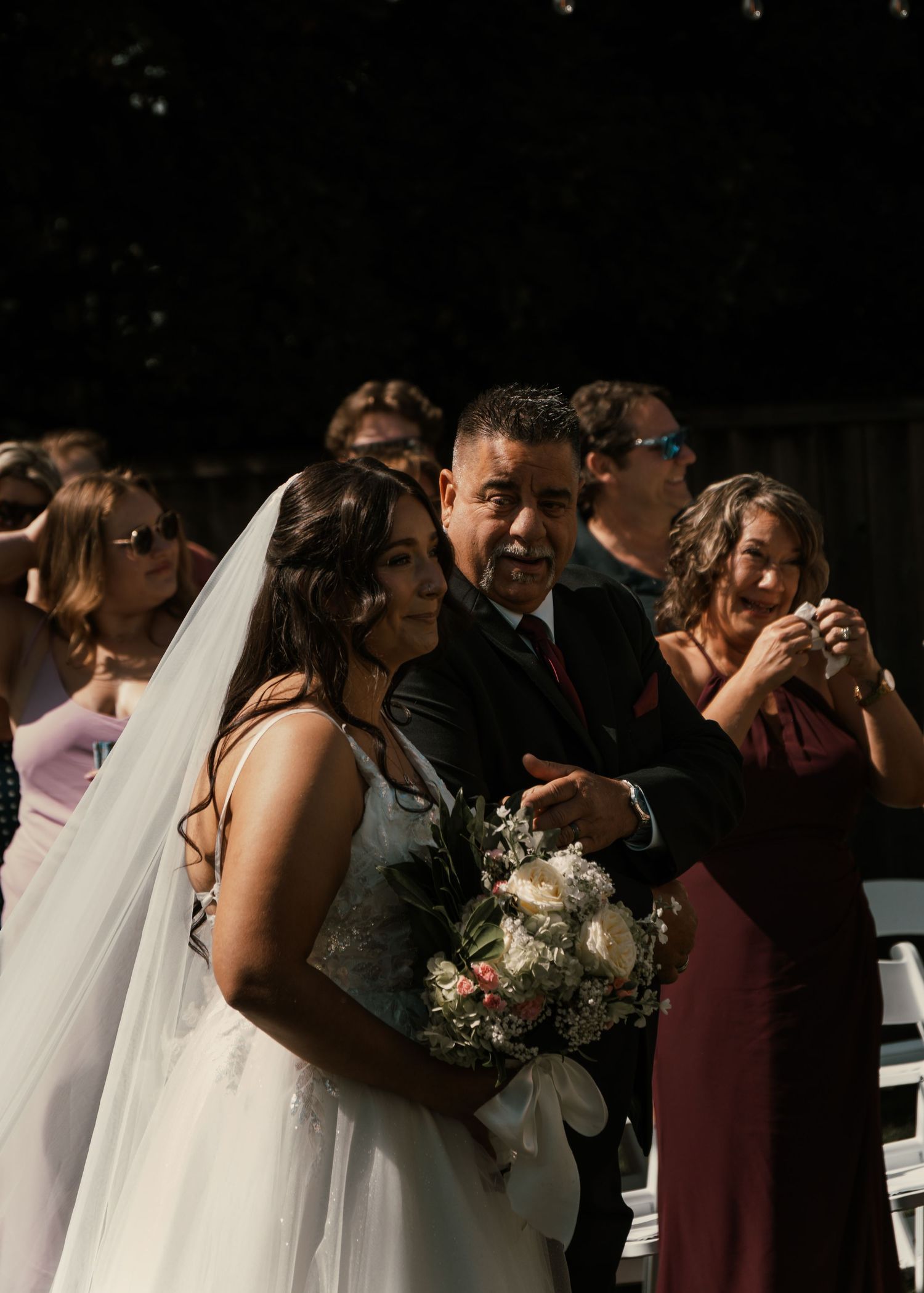 Bride in white gown and veil holds bouquet while walking with family at outdoor wedding ceremony.