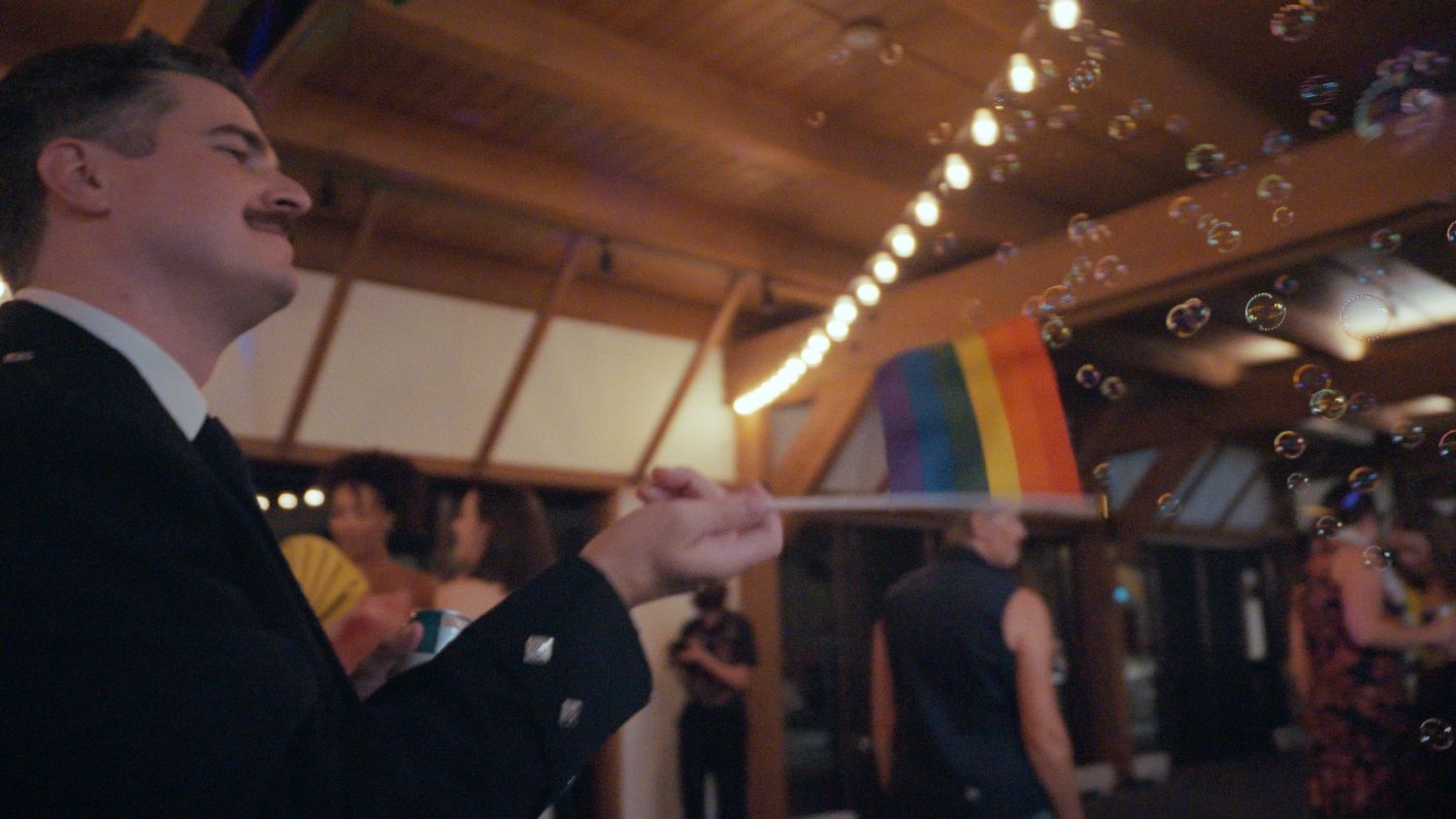 Wedding guest waving a rainbow pride flag inside a decorated reception hall with bubbles floating nearby.